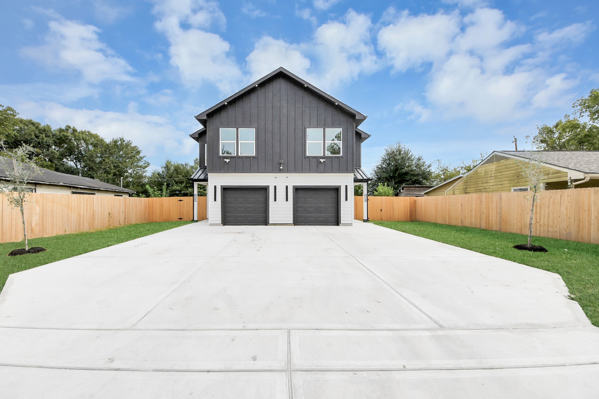 Modern two-story home with a striking dark facade, featuring dual garages and a spacious concrete driveway. The property is surrounded by a wooden privacy fence and landscaped with fresh greenery, offering a blend of style and functionality.