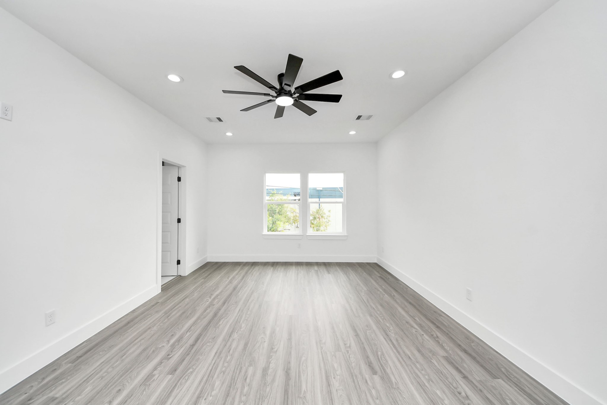 Spacious room with sleek wood flooring, fresh white walls, and a modern ceiling fan, offering ample natural light through a large window.