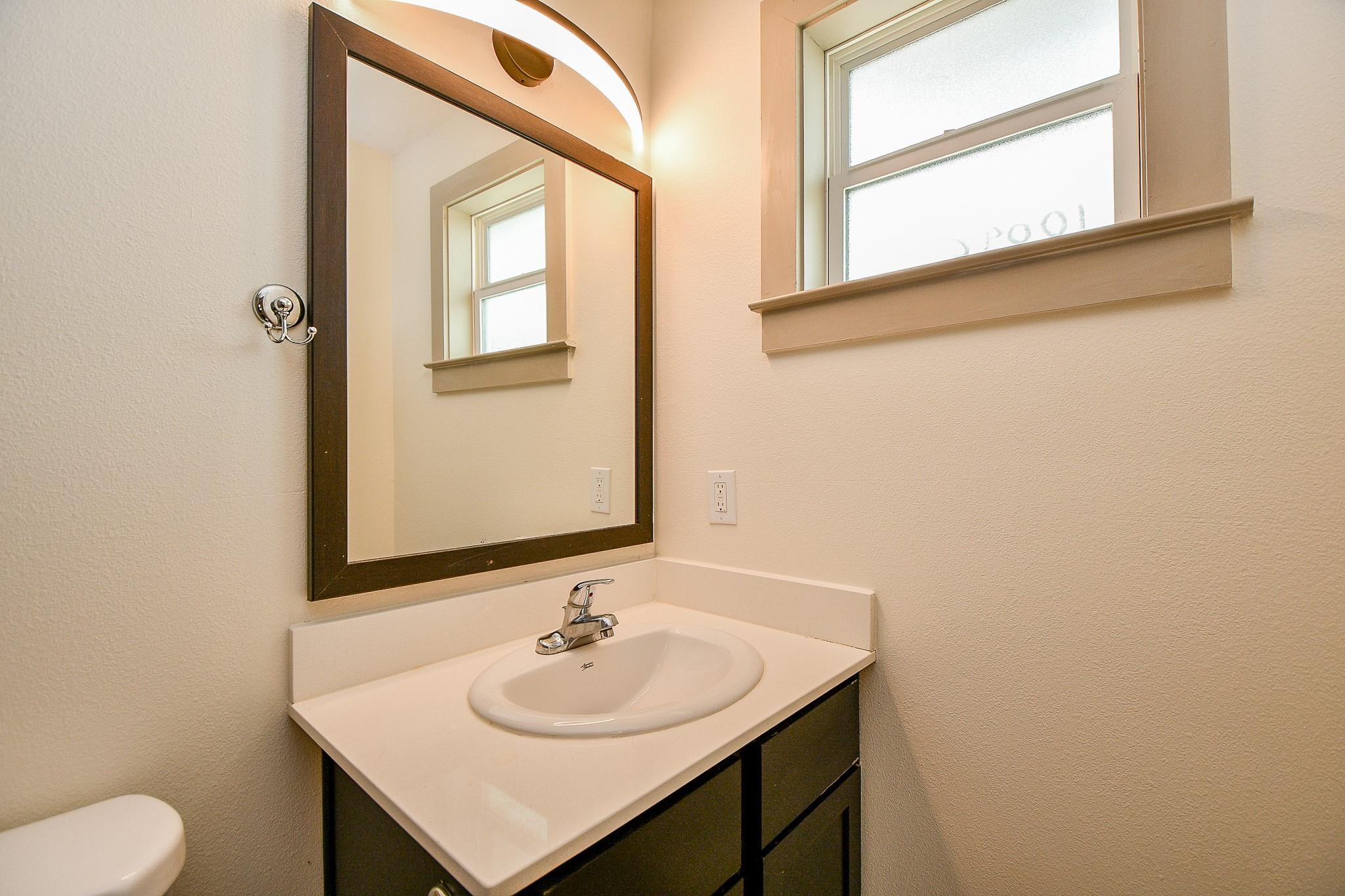 Single vanity with a sleek, dark cabinet. It has a large mirror with overhead lighting and a bright window providing natural light.