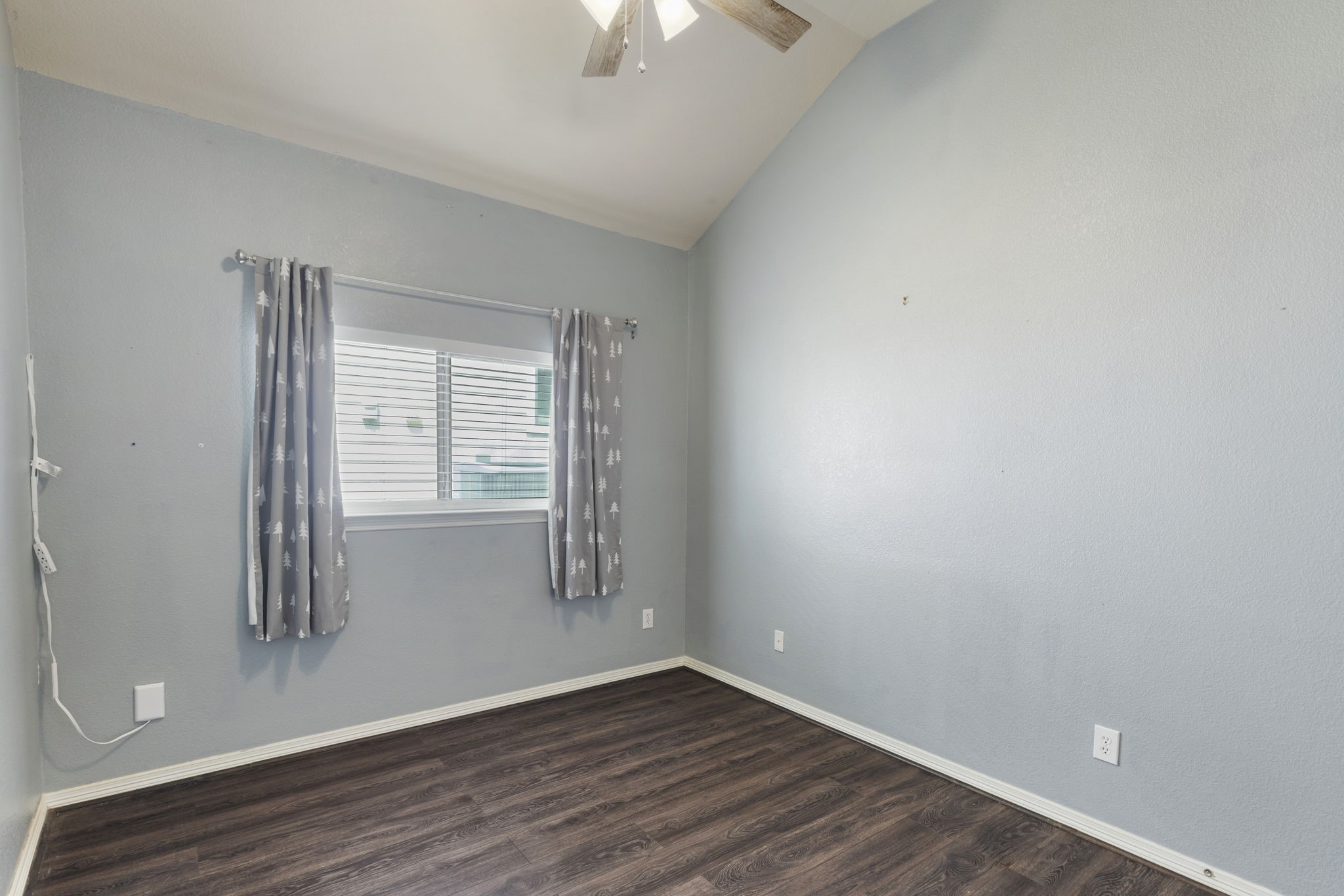 A bright room featuring a vaulted ceiling, laminate flooring, and a large window with curtains, perfect for a cozy bedroom or office space.