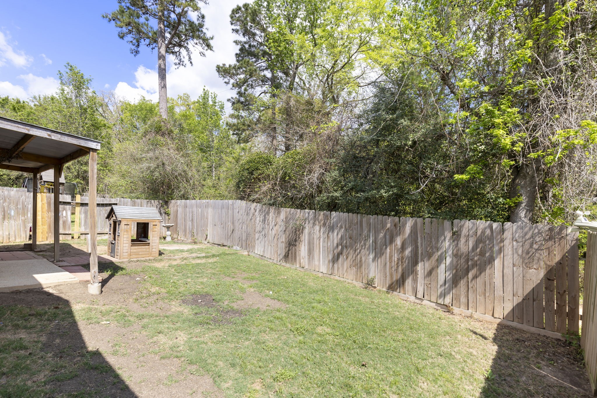 Charming backyard with a cozy covered patio and a quaint playhouse, bordered by lush greenery and a wooden fence, perfect for outdoor enjoyment.
