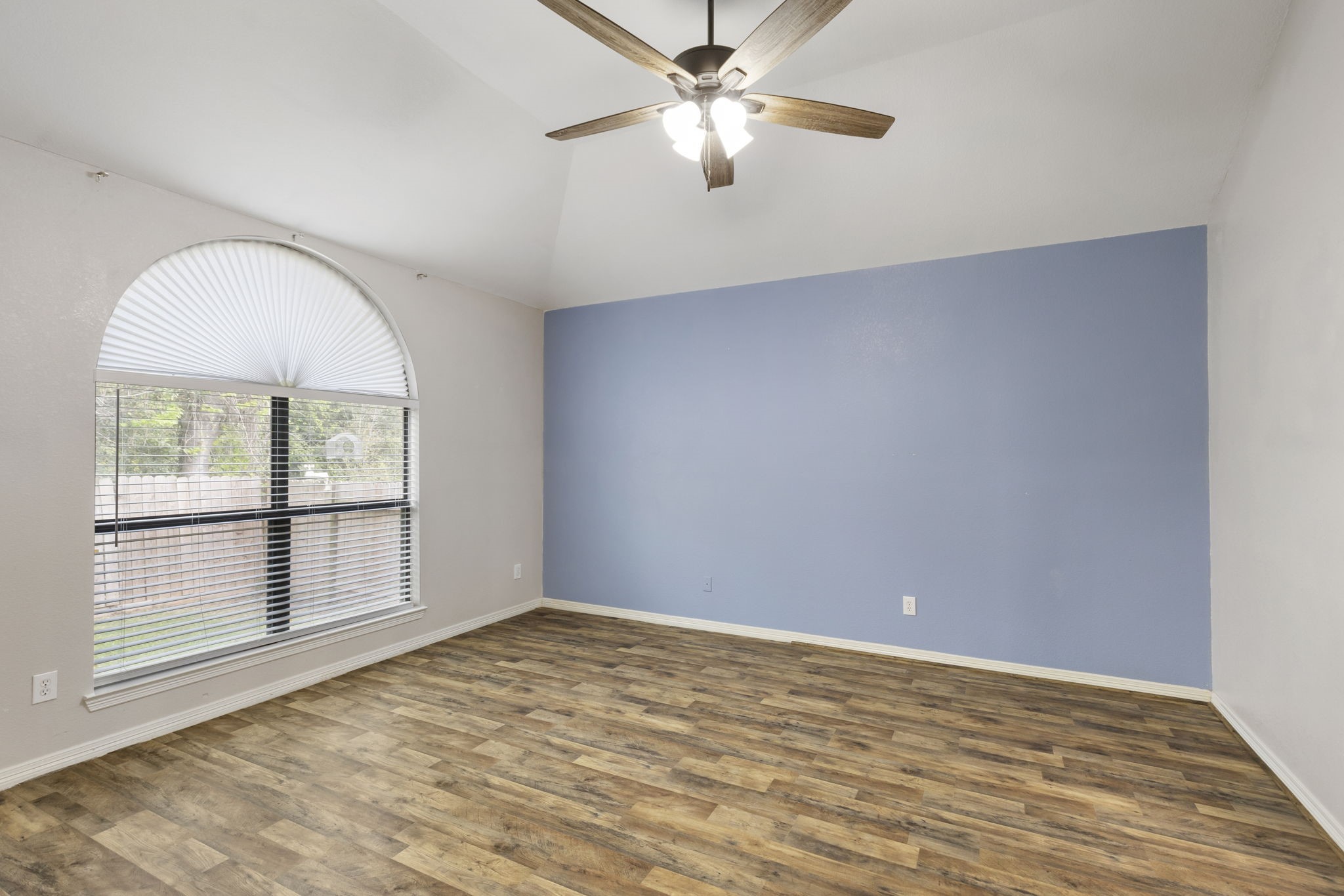 Experience tranquility in this spacious primary bedroom featuring a soothing blue accent wall, elegant arched window, and rich laminate flooring, perfect for restful nights.