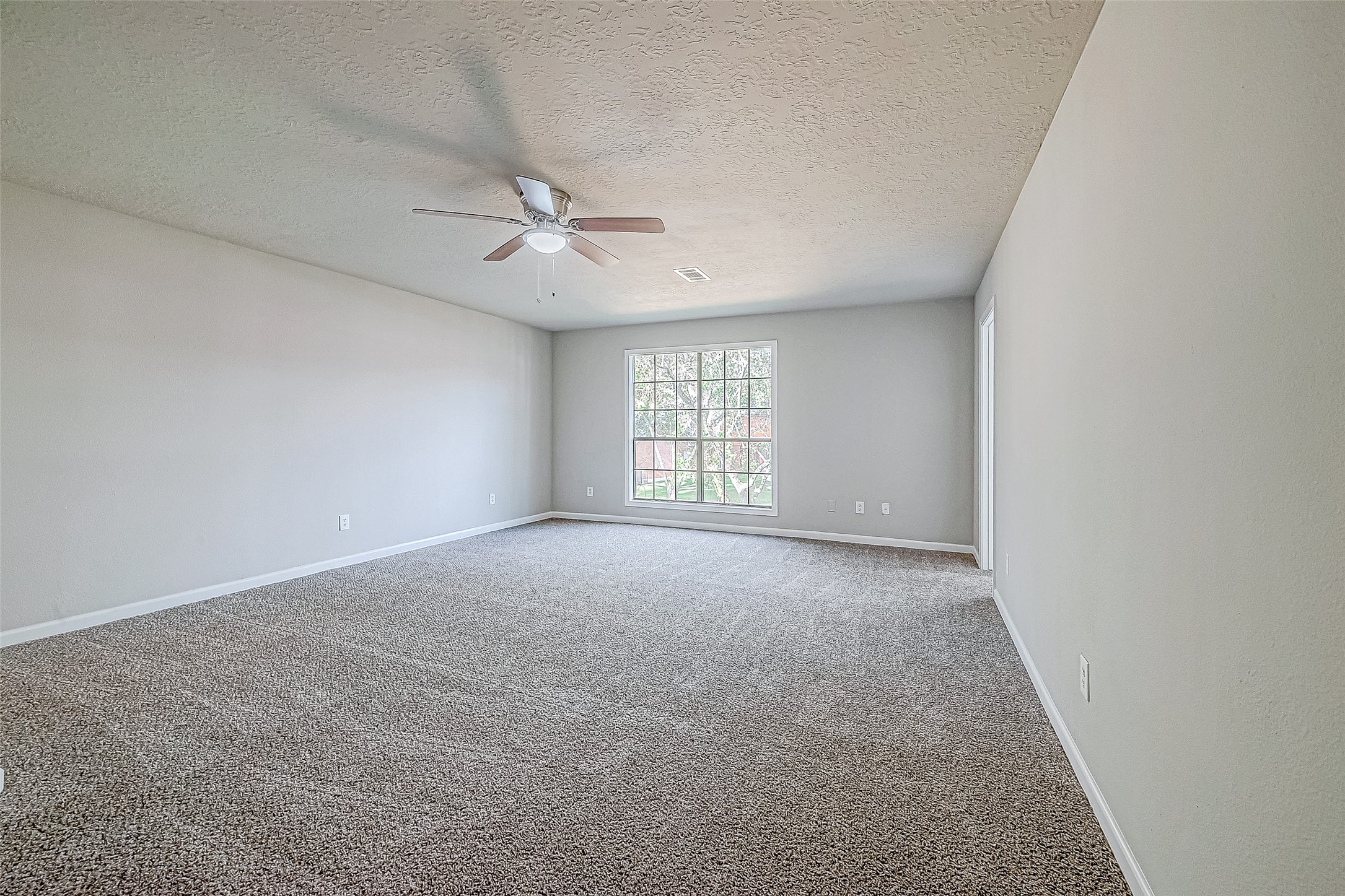 Spacious bedroom with neutral walls, carpeted flooring, large window for natural light, and a ceiling fan.