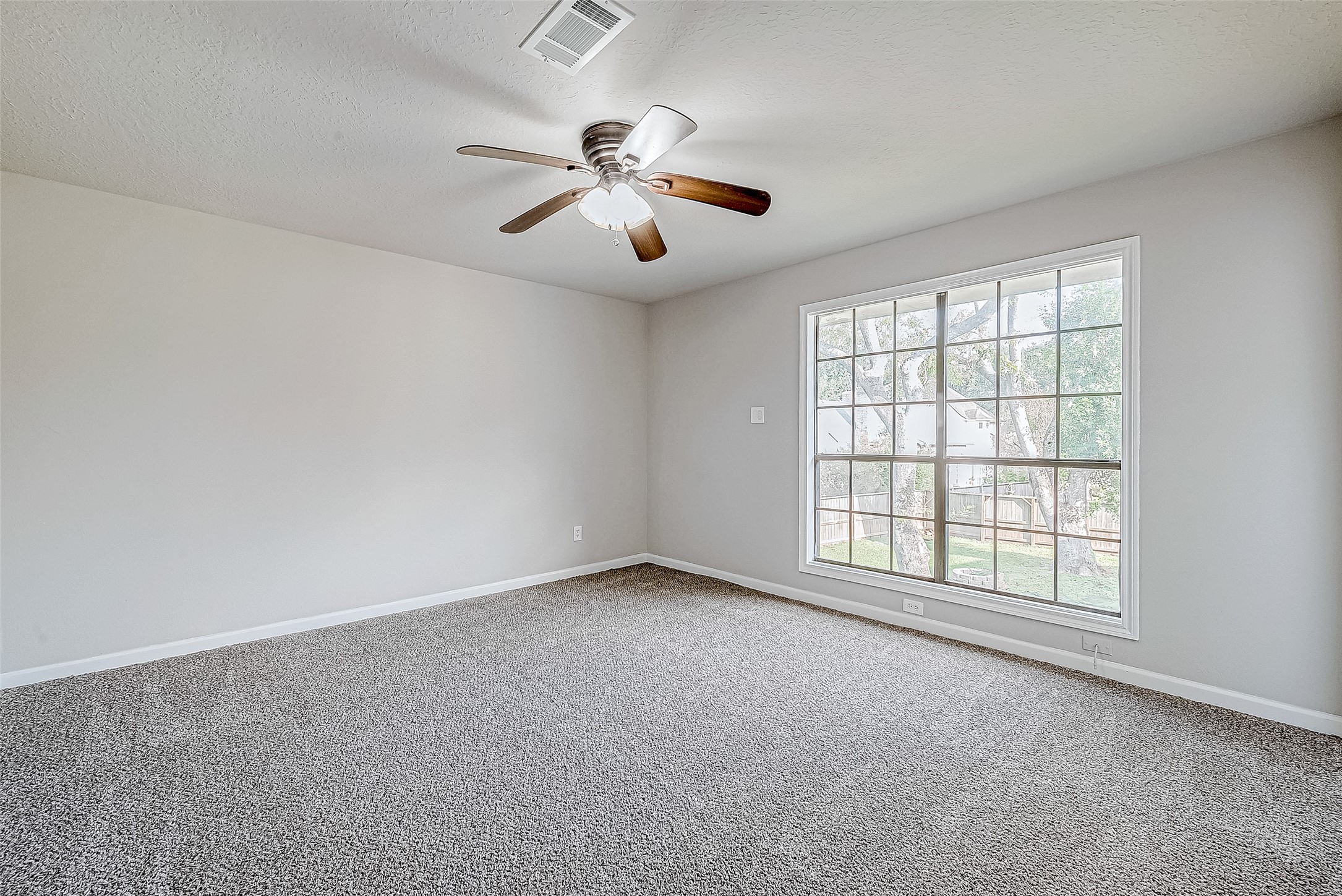 Bright and inviting bedroom with a large window overlooking the backyard, cozy carpet, and a ceiling fan with lights.
