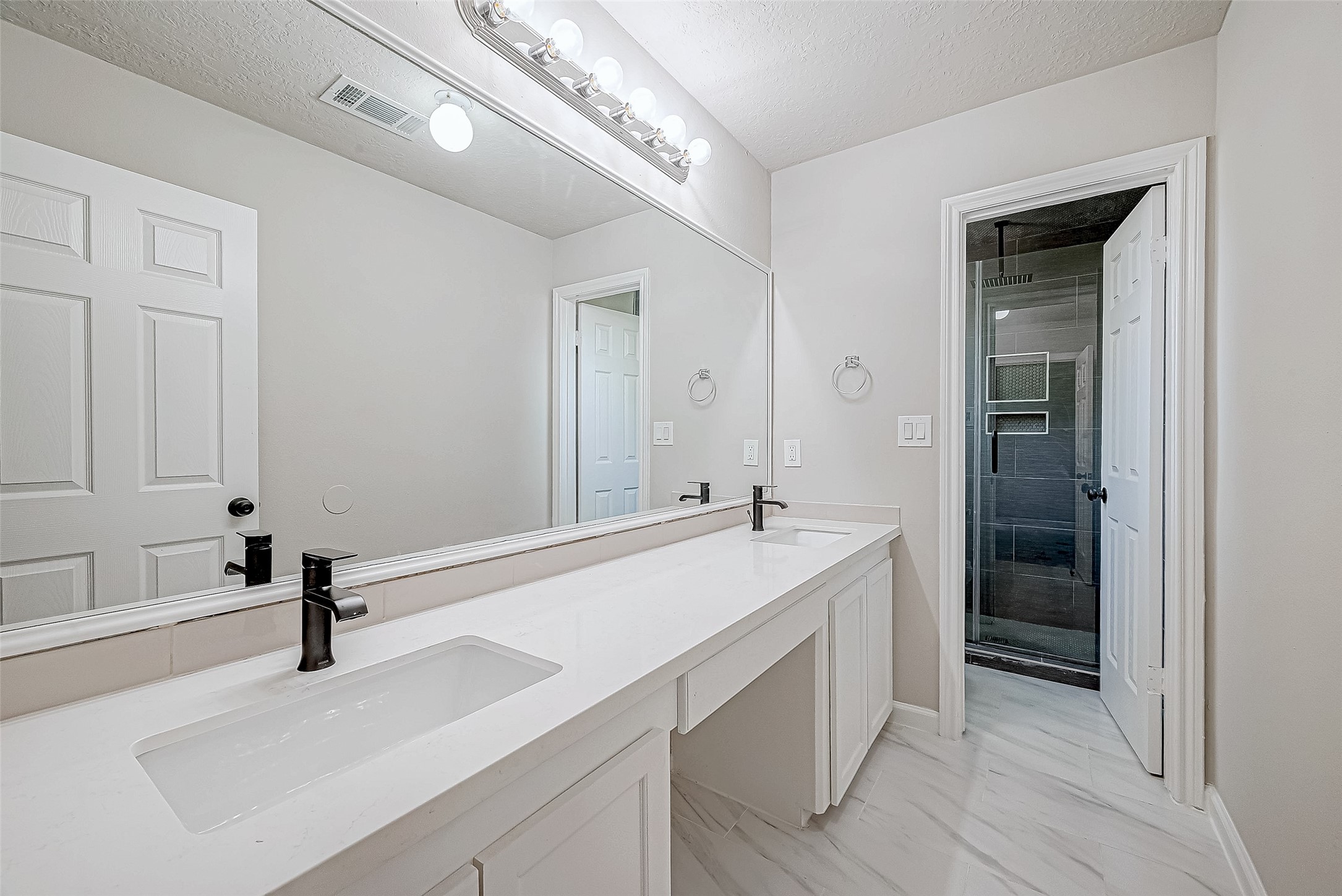 This bathroom features a double vanity with sleek black faucets, large mirror, and a stylish walk-in shower with dark tiling, creating a clean and contemporary look.