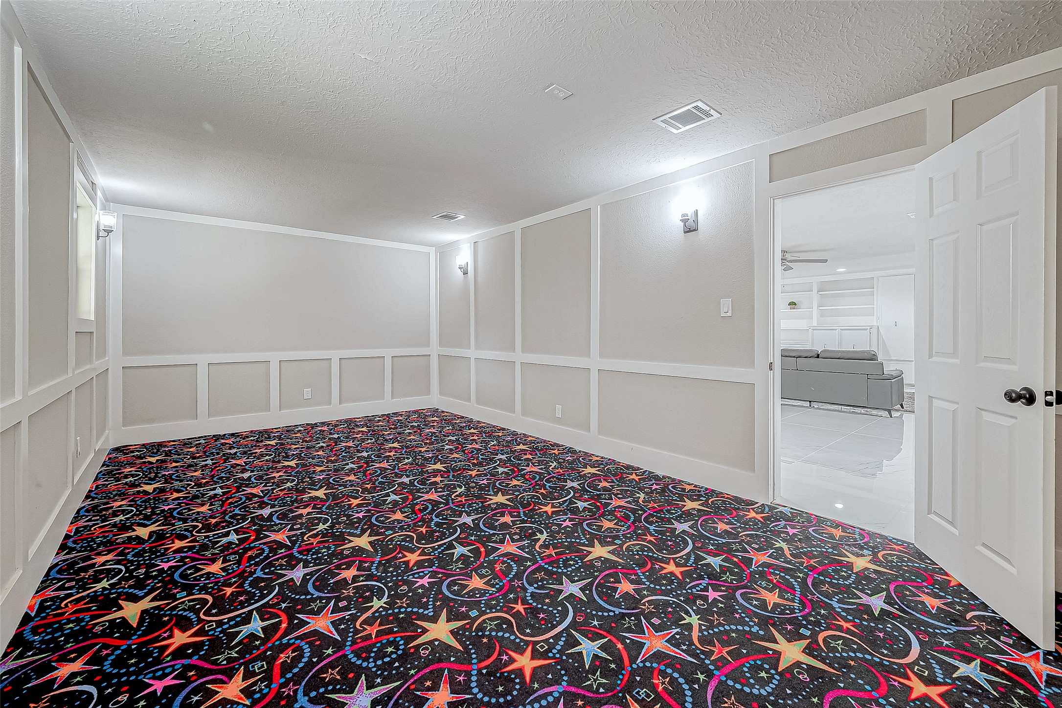 The versatile room features a vibrant, star-patterned carpet. An open doorway connects to the living area, perfect for a home theater or playroom.