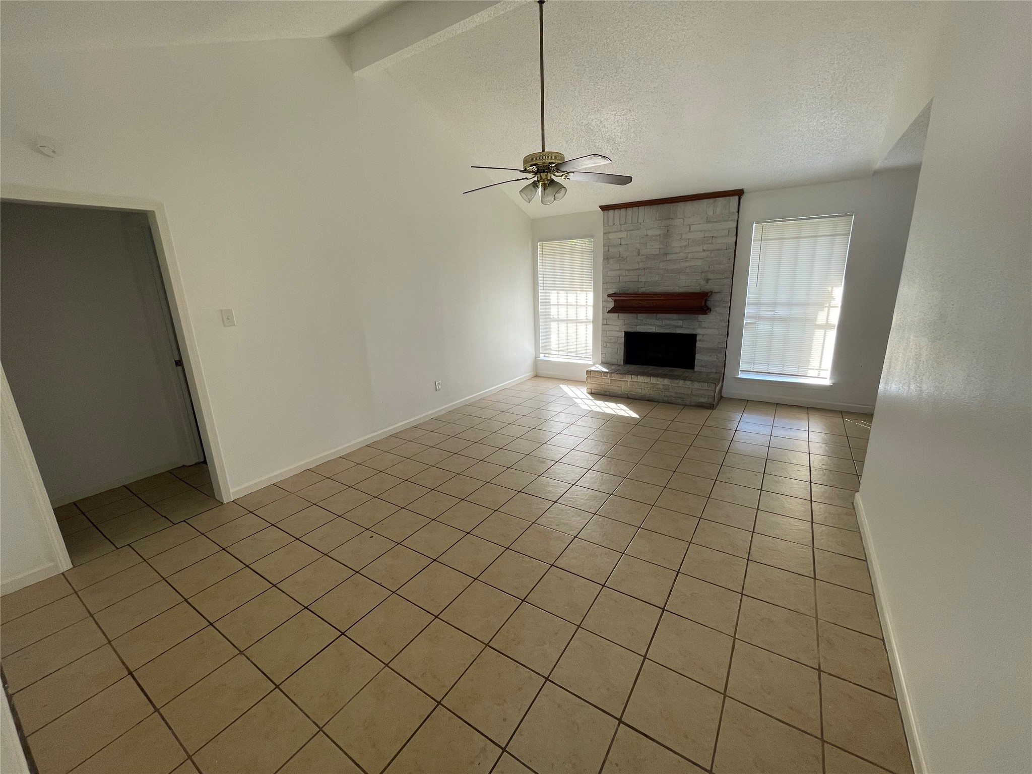 Spacious living area featuring a vaulted ceiling, neutral paint, ceramic tile flooring, and a ceiling fan. A whitewashed brick fireplace with a wood mantel serves as the focal point, while two large windows provide natural light.