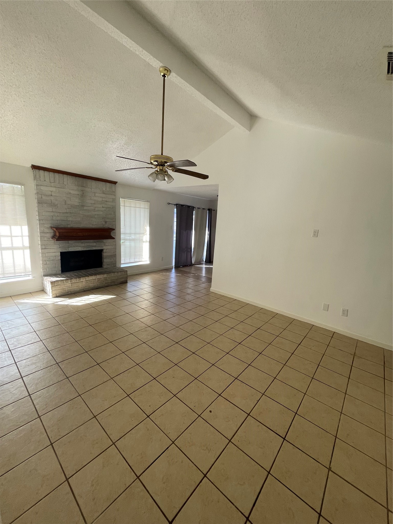 Alternate view of the spacious living area featuring vaulted ceilings, ceramic tile flooring, neutral walls, and a ceiling fan. The whitewashed brick fireplace with a wood mantel adds character, while large windows provide natural light. The open layout offers flexibility for furniture placement and entertaining.