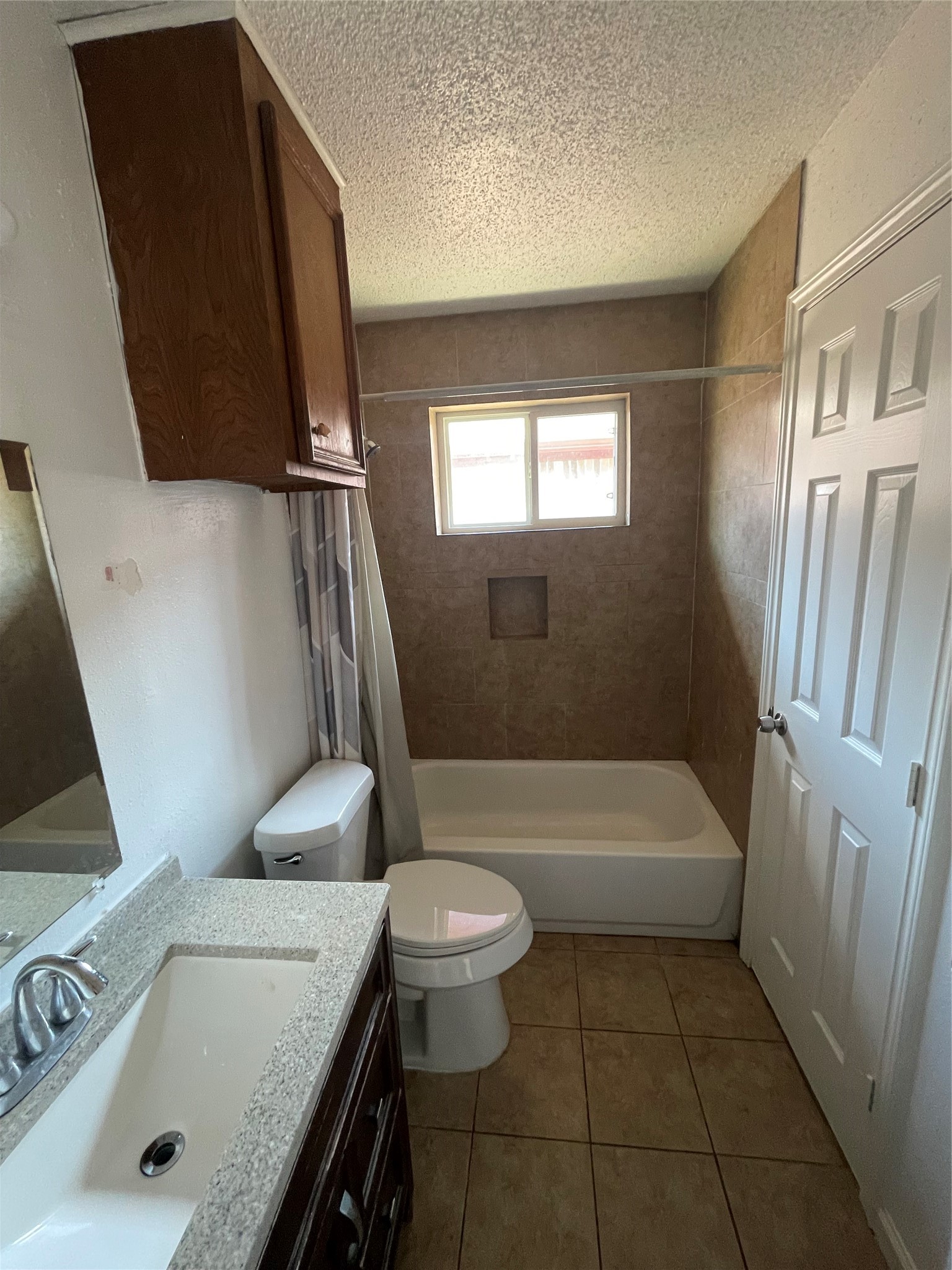 Full bathroom featuring a tub and shower combination with tiled surround, built-in shower niche, and window providing natural light. Additional features include a vanity with countertop sink, wood cabinetry, overhead storage, and tile flooring.