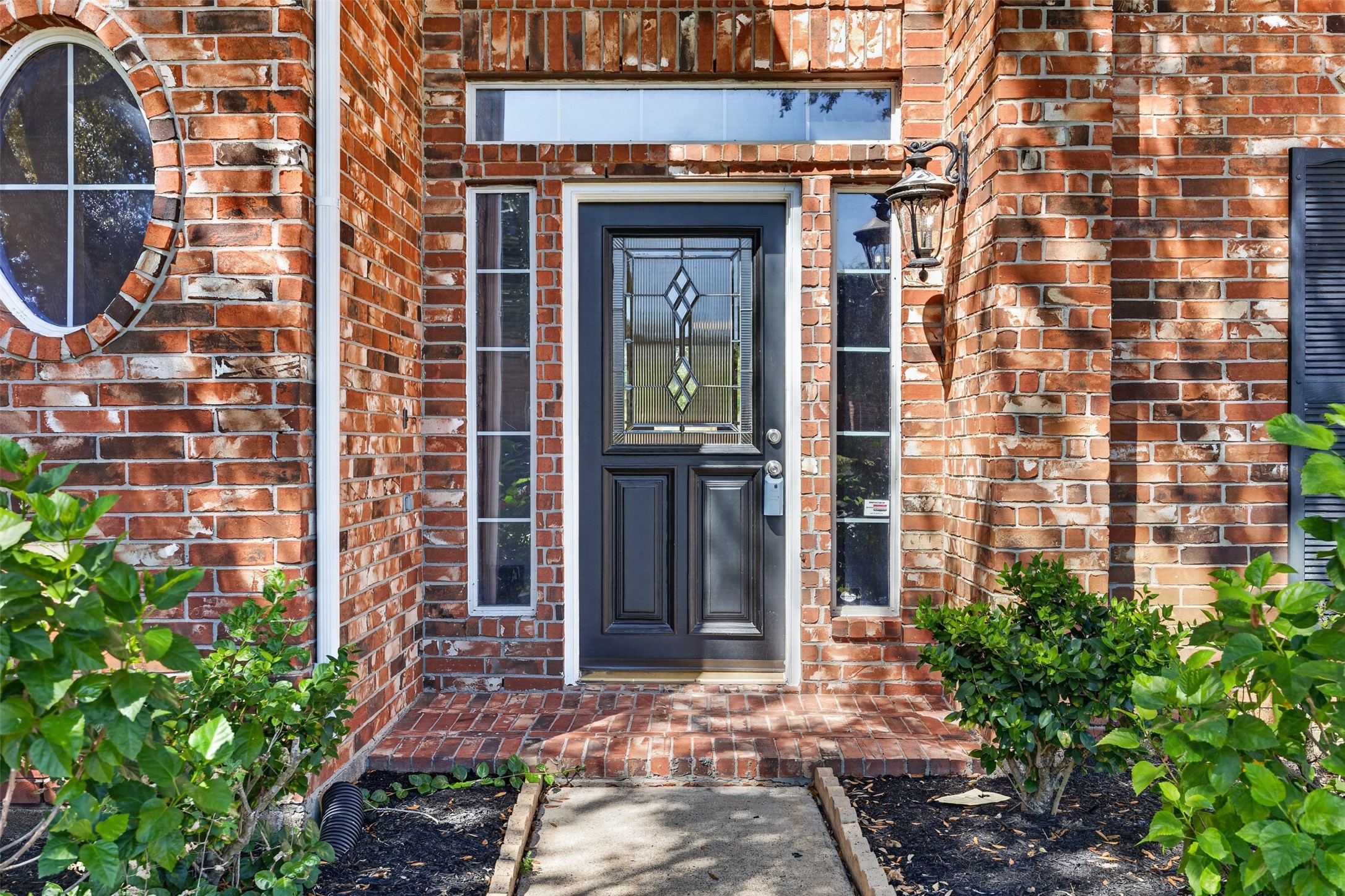 Charming brick entryway highlighted by a stylish front door with decorative glass panels, framed by lush greenery and elegant lighting.