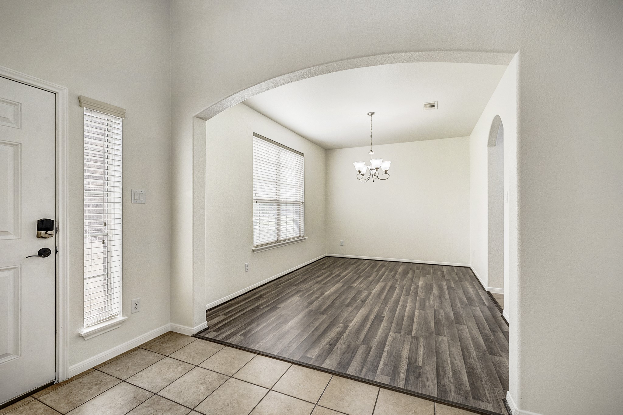 Bright and inviting entryway featuring elegant tile flooring transitioning to wood-look floors, with an arched opening leading to a spacious dining area enhanced by a charming chandelier.
