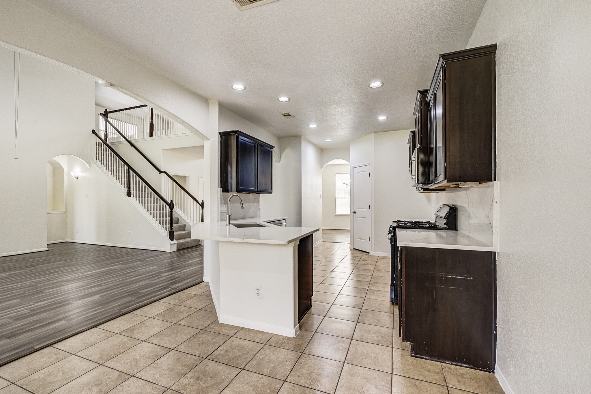 Bright and open kitchen with dark cabinetry and modern fixtures, seamlessly connected to a spacious living area with elegant flooring and a stylish staircase.