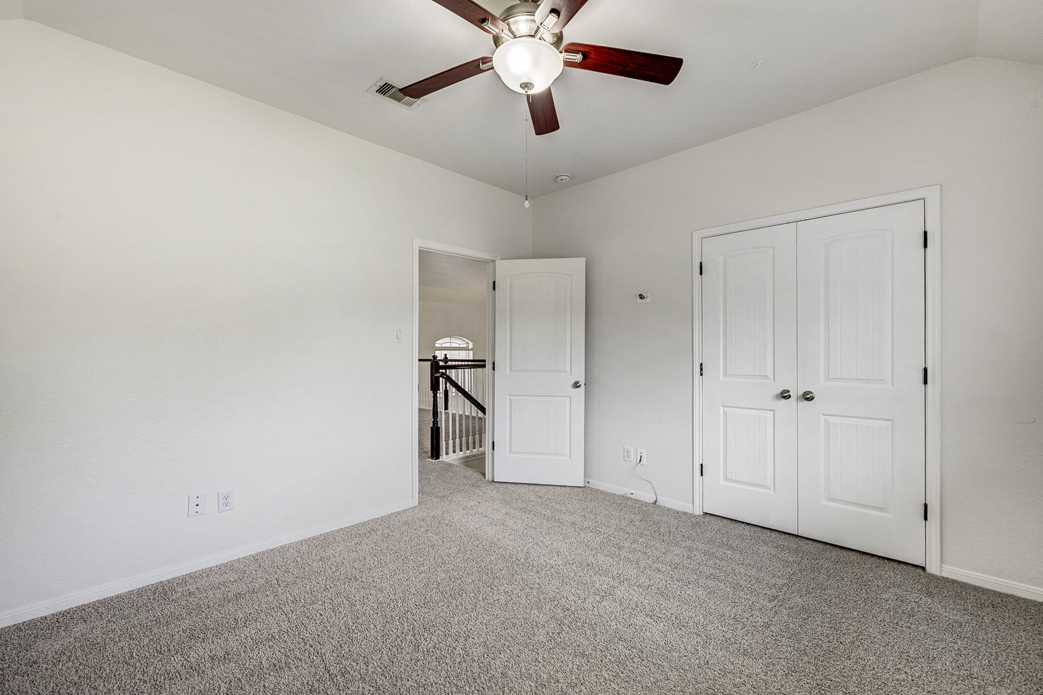 Spacious room with soft carpeting, neutral walls, and a ceiling fan, featuring a double-door closet and an open doorway leading to a hallway with elegant railings.