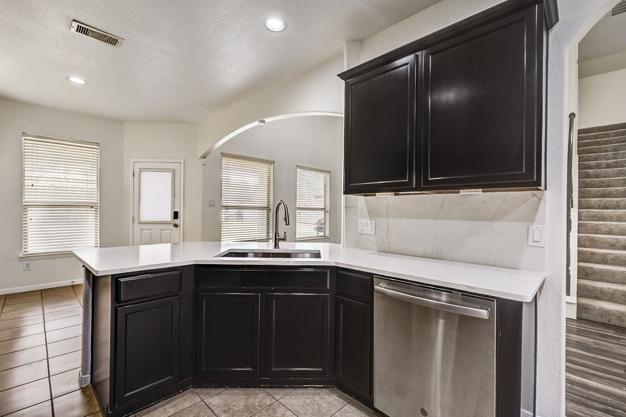 Elegant kitchen featuring dark cabinetry, a sleek stainless steel dishwasher, and a spacious countertop with a modern sink, complemented by abundant natural light from large windows and convenient access to the stairway.