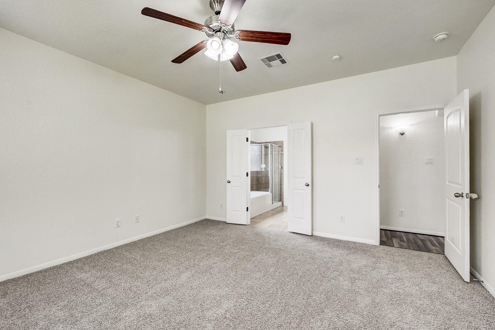 Spacious bedroom featuring plush carpeting, a ceiling fan, and direct access to a modern bathroom with double doors.