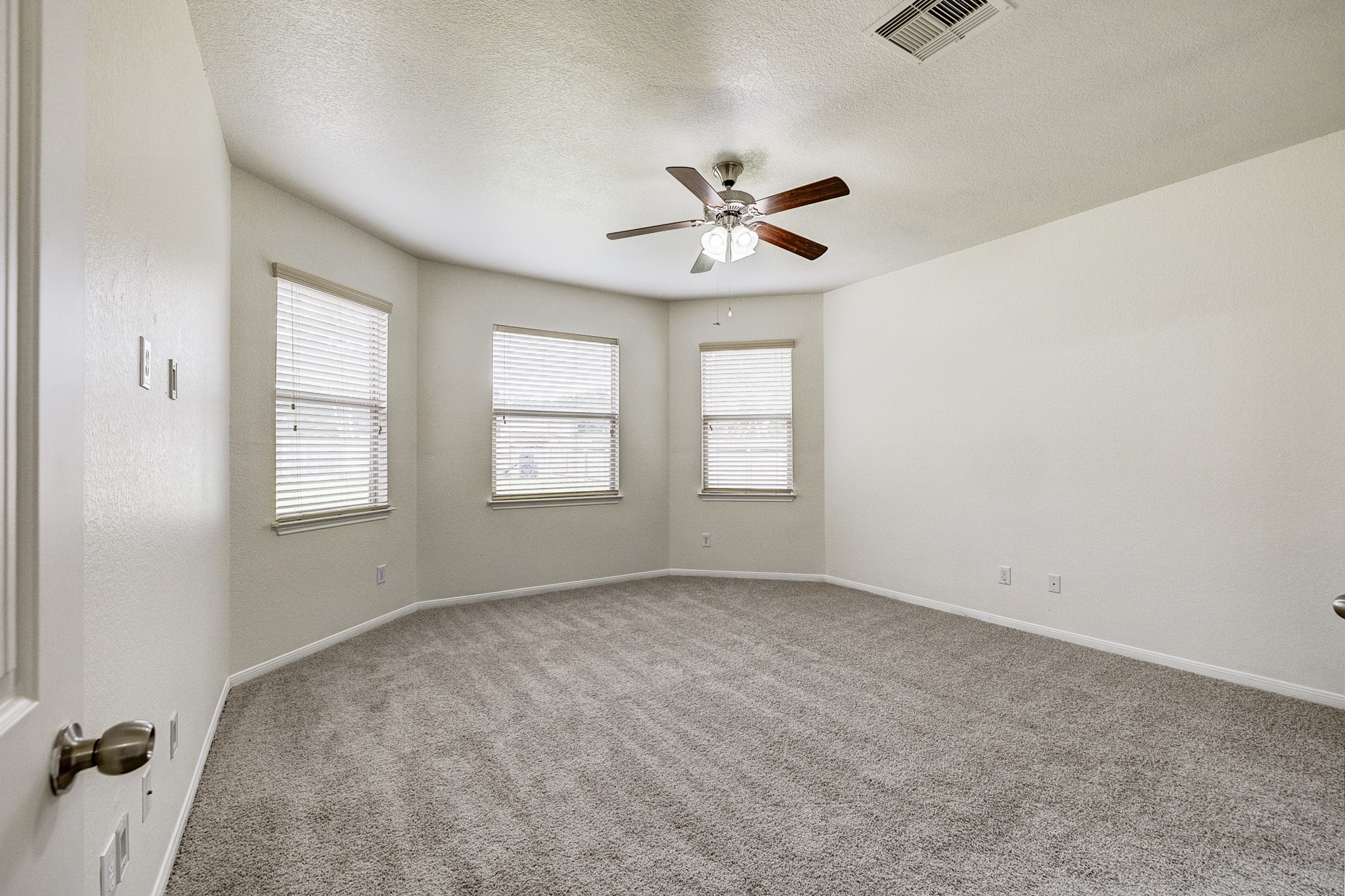 Spacious bedroom featuring neutral-toned carpet, three large windows for natural light, and a modern ceiling fan.