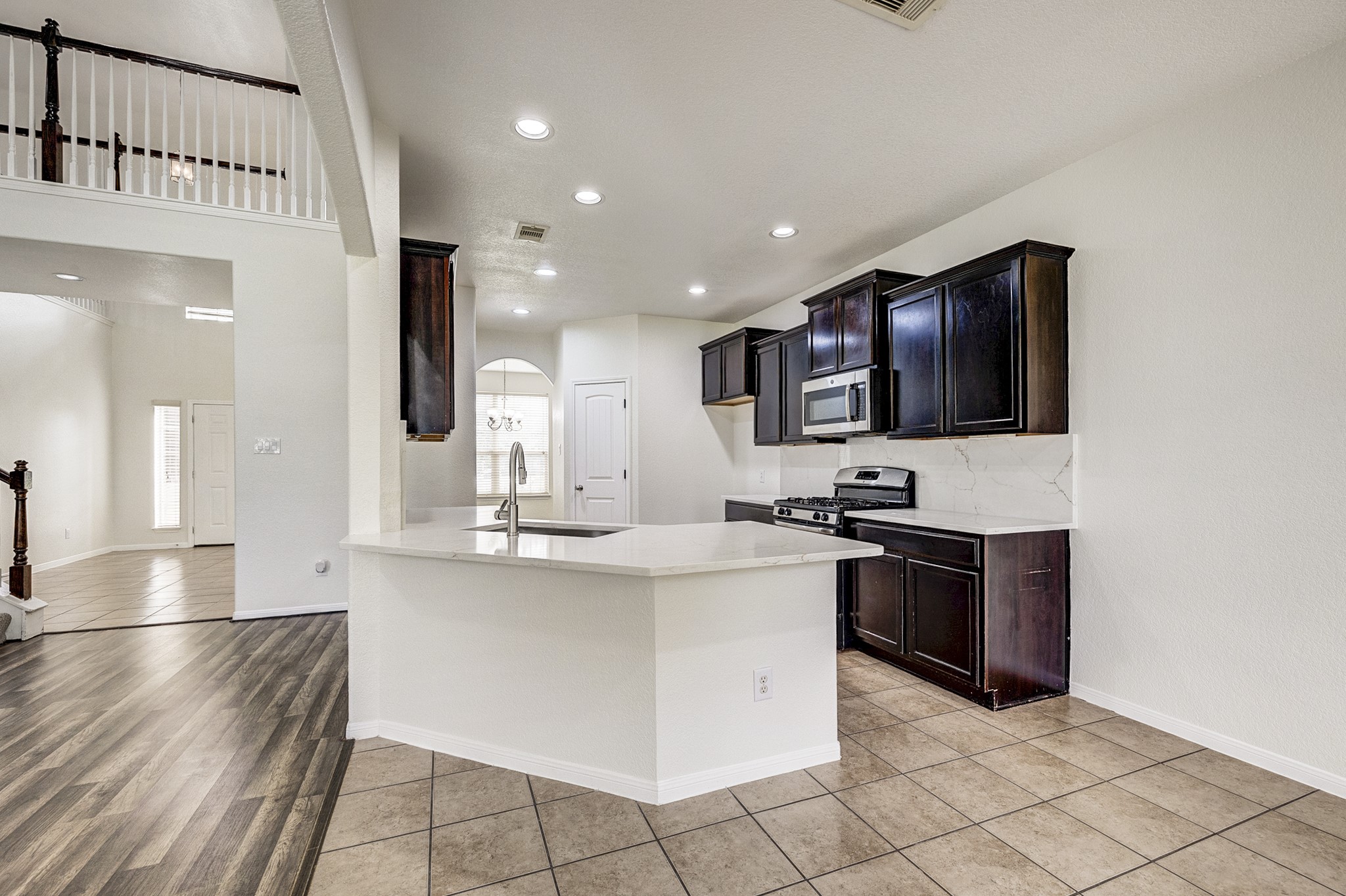 Modern kitchen with sleek dark cabinetry, stainless steel appliances, and a spacious white countertop. Open layout leads to a bright entryway with elegant flooring and high ceilings.