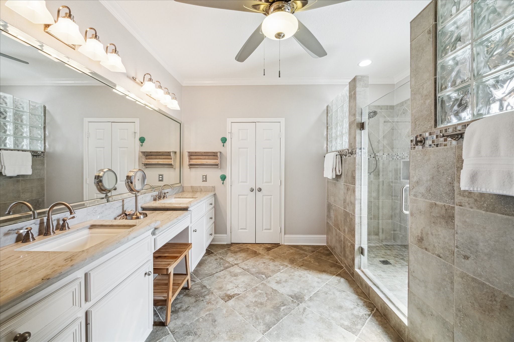 The primary  bathroom features a spacious layout with a double vanity, large mirrors, and ample lighting. It includes a double walk-in shower with glass block accents and neutral tile flooring. A ceiling fan adds comfort, and theres a sleek, modern design throughout.