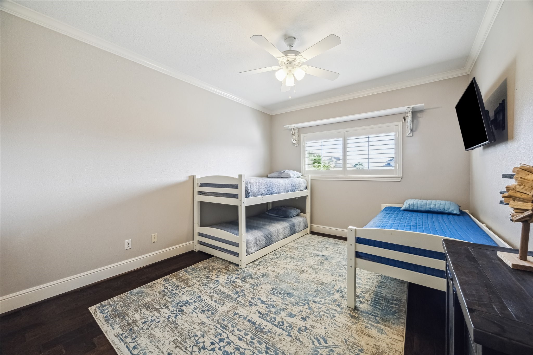 This secondary bedroom features a clean, modern design with neutral walls, dark hardwood floors, and a ceiling fan. It includes a bunk bed and a single bed, with a large window for natural light. A wall-mounted TV offers entertainment, and a patterned area rug adds warmth and style.