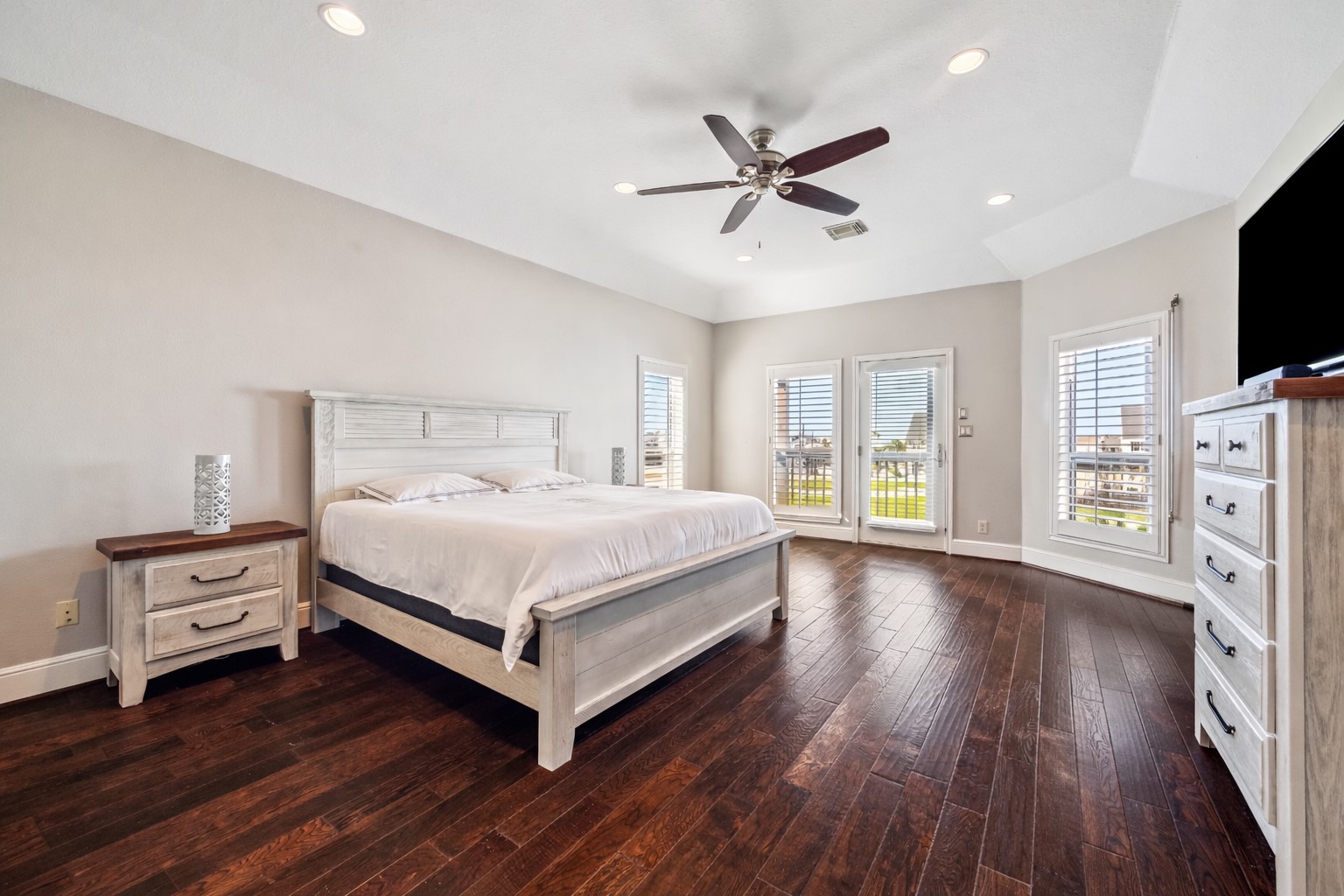 Spacious bedroom with hardwood floors and ample natural light from multiple windows. Ceiling fan, neutral walls, and elegant furniture, offering a comfortable and inviting atmosphere.