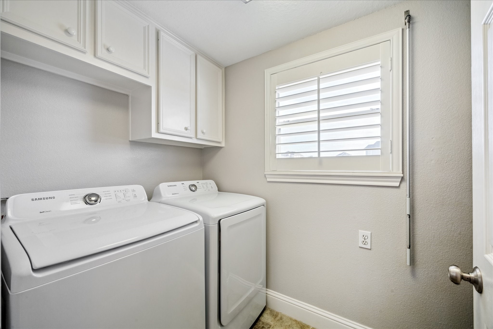 This laundry room is bright and functional, featuring a washer and dryer set, white cabinetry for storage, and a window with shutters for natural light.