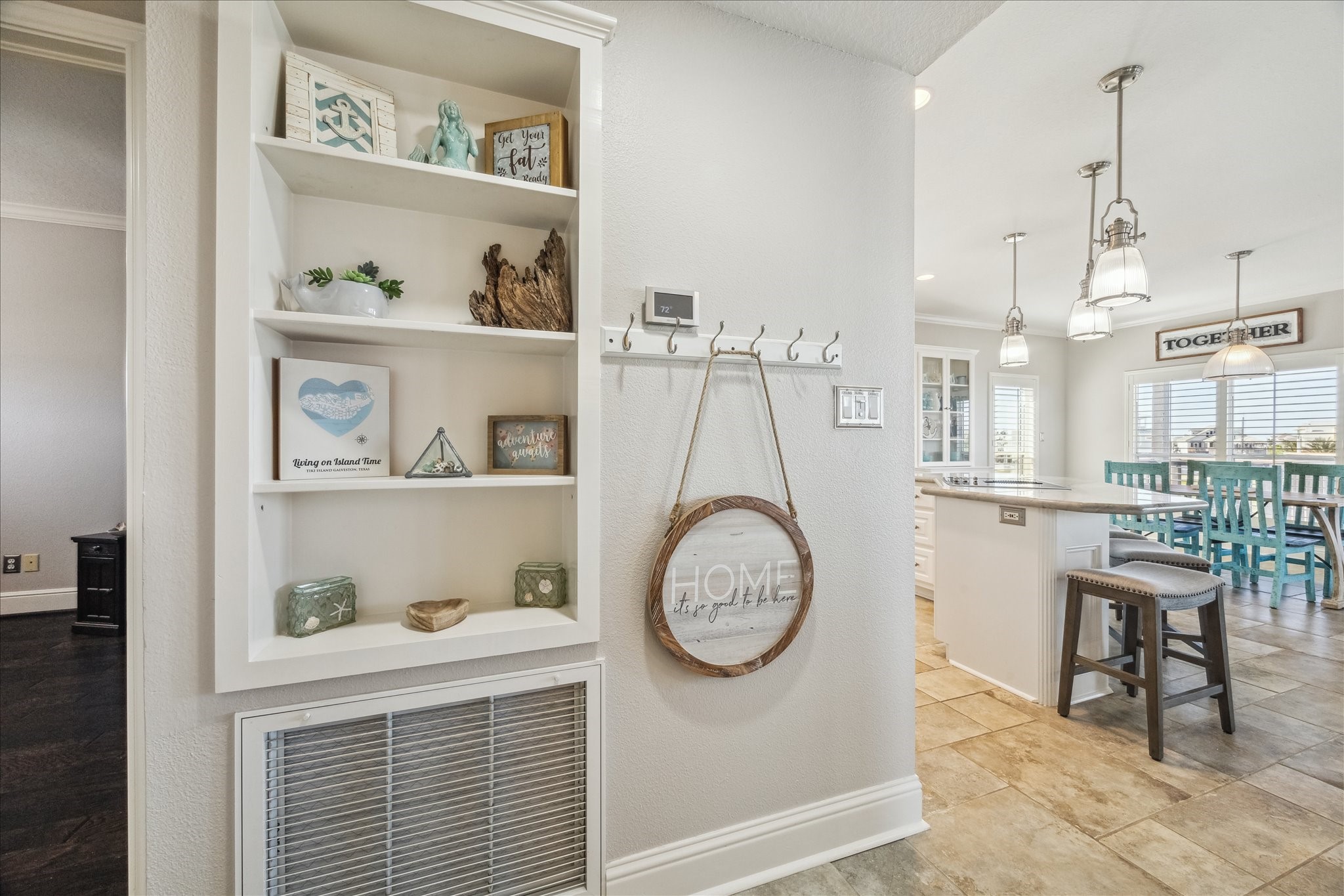 Bright, inviting interior with built-in shelving and decorative accents. The open layout leads to a stylish kitchen with pendant lighting, an island with seating, and a dining area with large windows. The space is modern and welcoming.