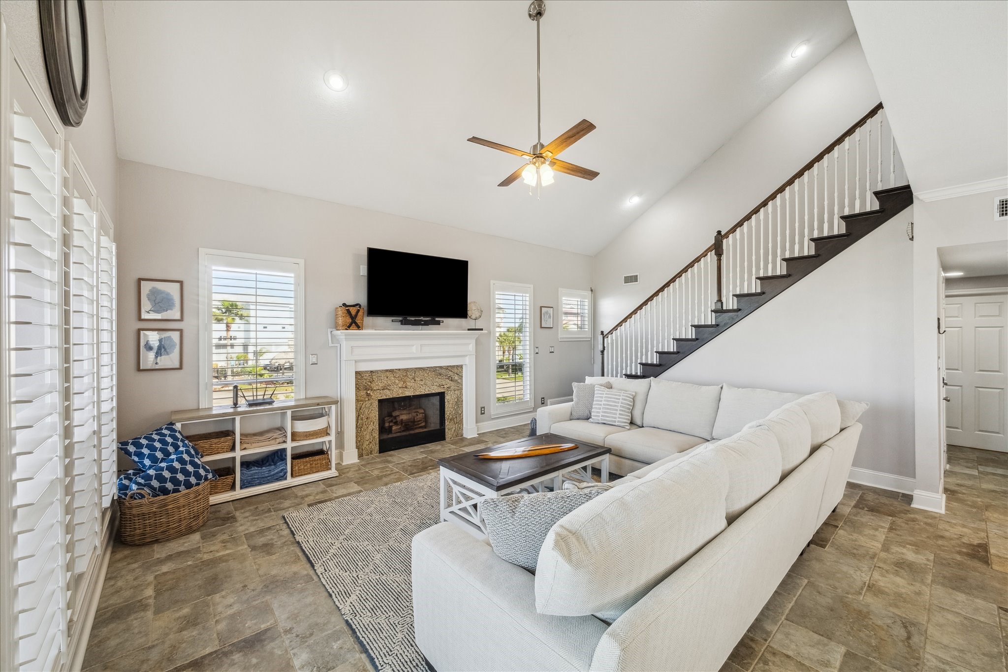 Bright and spacious living room with high ceilings, featuring a cozy fireplace, large sectional sofa, and a mounted TV. The room is well-lit with natural light from multiple windows, and a staircase leads to the upper level, enhancing the open feel.