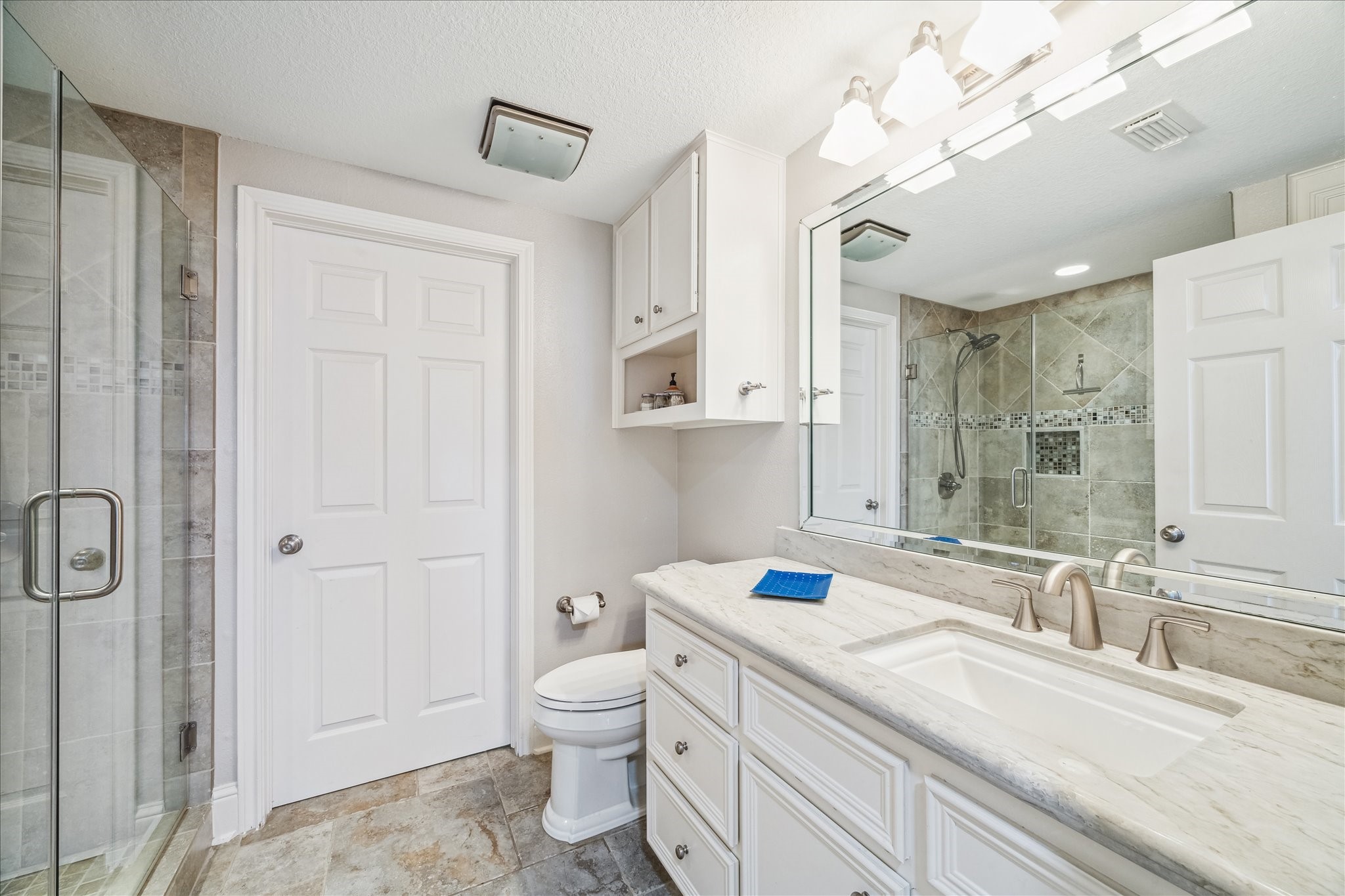 This secondary bathroom features a modern design with a large glass-enclosed shower, elegant tile work, a marble countertop with dual sinks, and ample cabinet storage. Bright lighting and neutral tones create a fresh and inviting atmosphere.