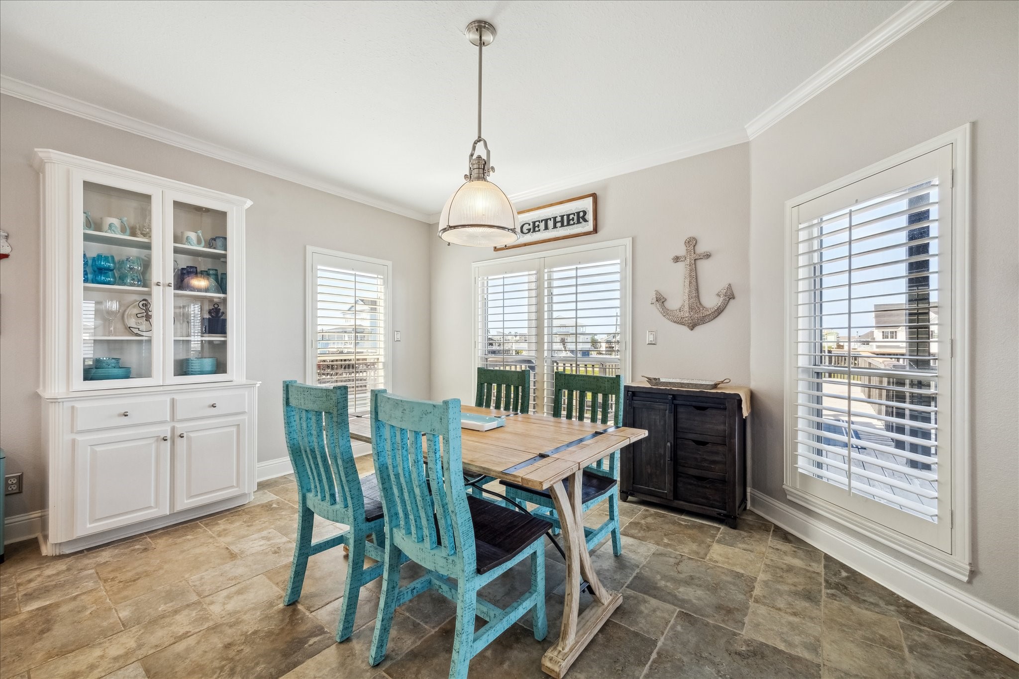 Bright dining area with a coastal vibe, featuring a wooden table, turquoise chairs, and a white cabinet. Large windows with shutters allow ample natural light, while nautical decor adds charm. Perfect for casual dining and entertaining.