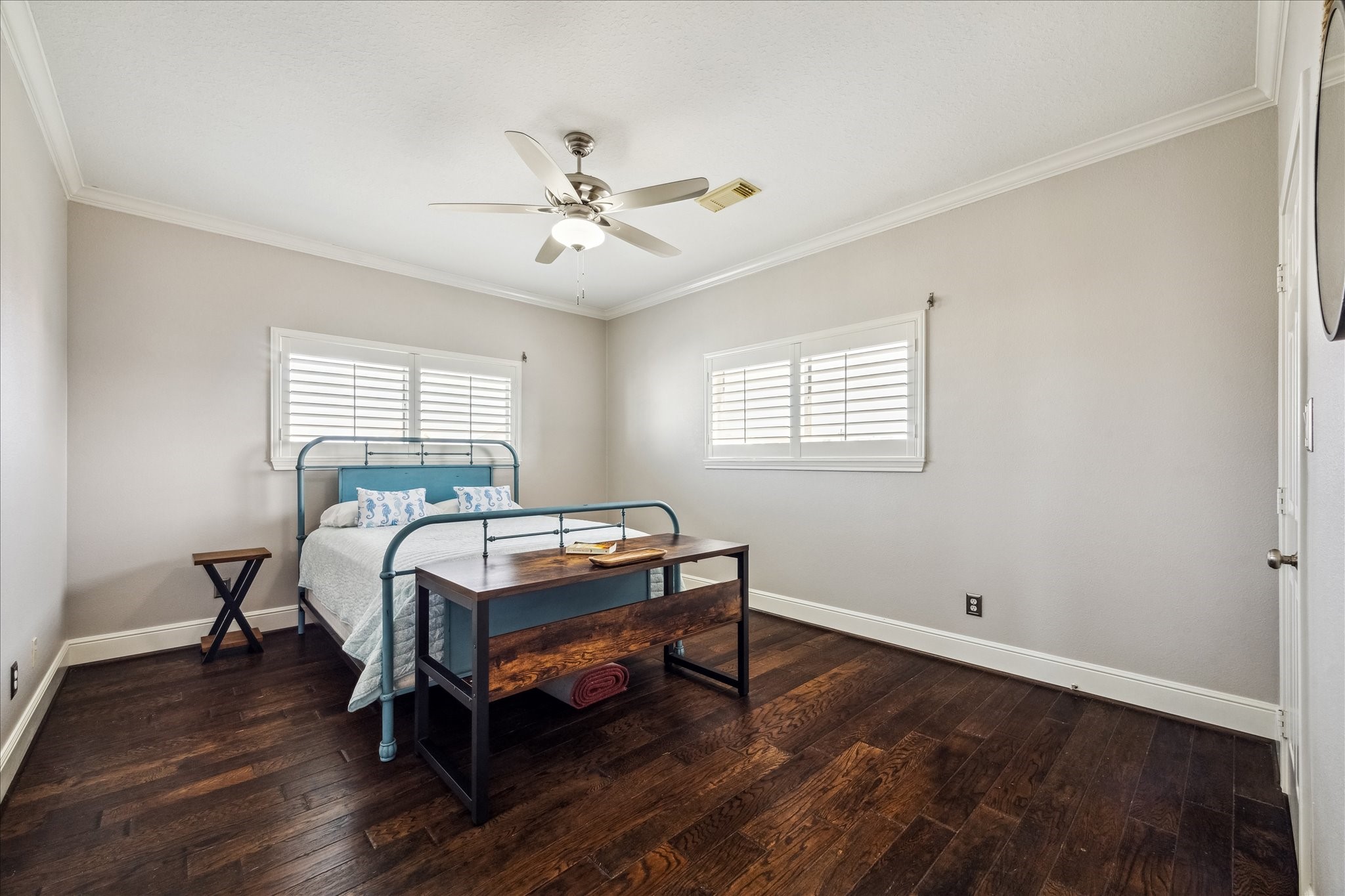This secondary bright bedroom features hardwood floors, neutral walls, and a ceiling fan. It includes a bed with a wooden bench at the foot and ample natural light from two windows with plantation shutters. The closet could be converted to a 3rd bath , plumbing is accessible.