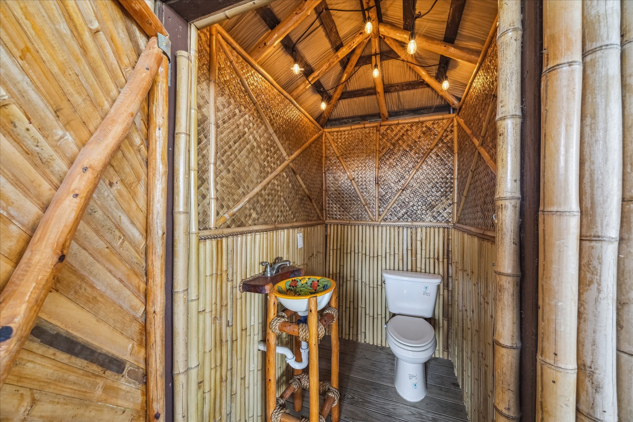 This is a unique, rustic bathroom featuring bamboo walls and ceiling, with a colorful, artistic sink and modern toilet. Warm lighting adds a cozy ambiance, perfect for a tropical or nature-inspired home. Not permittable or insurable.