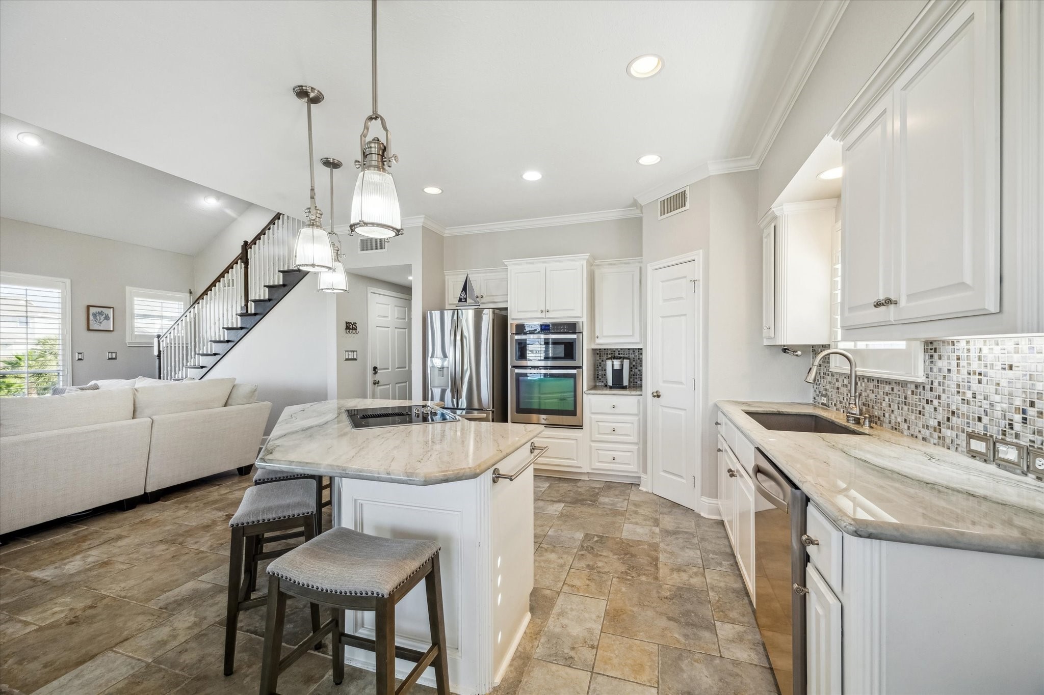 Bright, open-concept kitchen and living area. The kitchen features modern stainless steel appliances, white cabinetry, and a stylish tile backsplash. A central island offers additional seating and workspace. Adjacent is a cozy living area with a staircase leading upstairs, creating a welcoming and functional space.