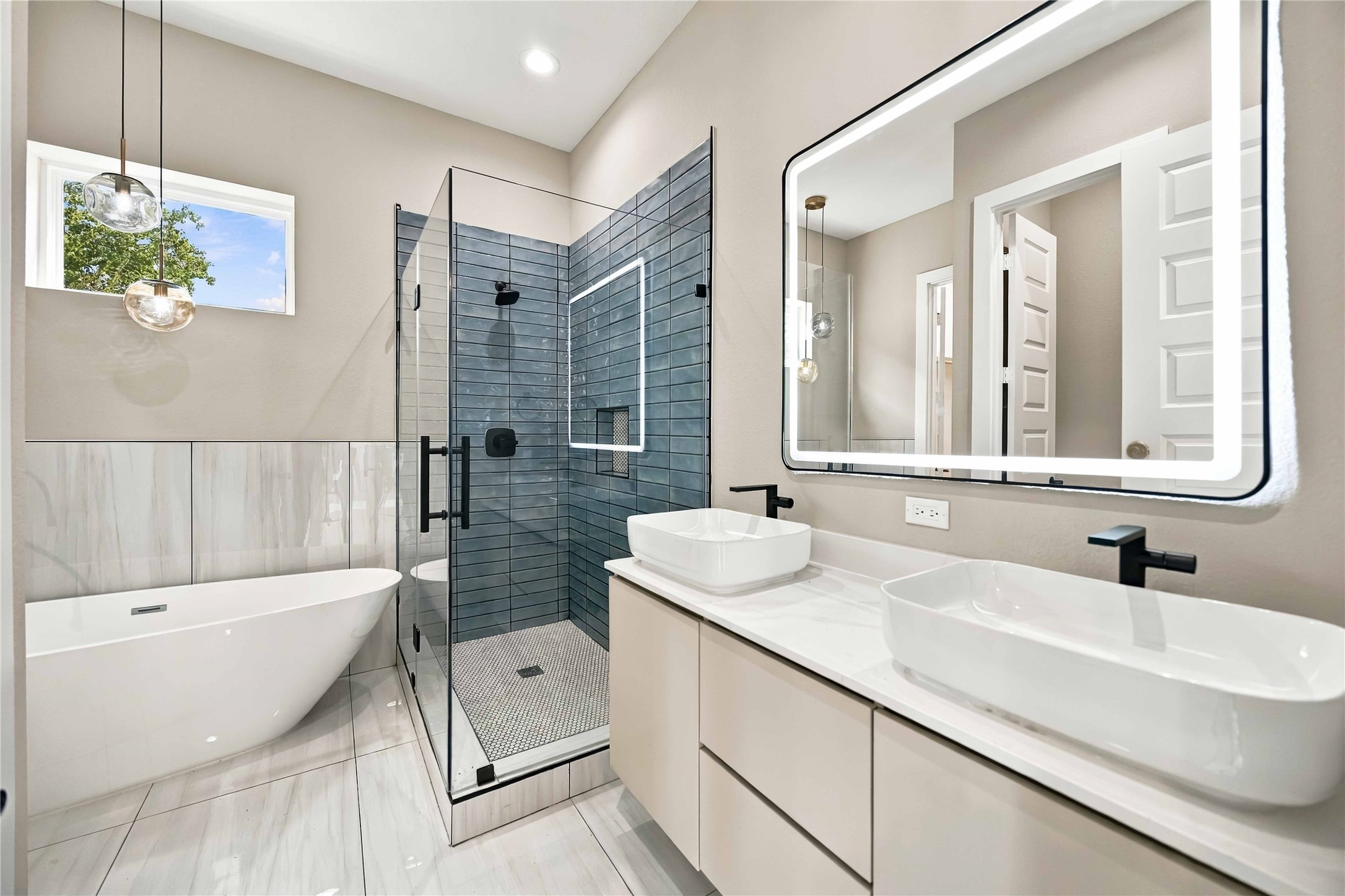 Spa-inspired primary bathroom featuring a freestanding soaking tub, glass-enclosed walk-in shower with contemporary blue-gray tile, dual vessel sinks, a sleek white vanity with light countertops, and a striking illuminated mirror. Matte black fixtures and elegant pendant lighting add a sophisticated modern finish.