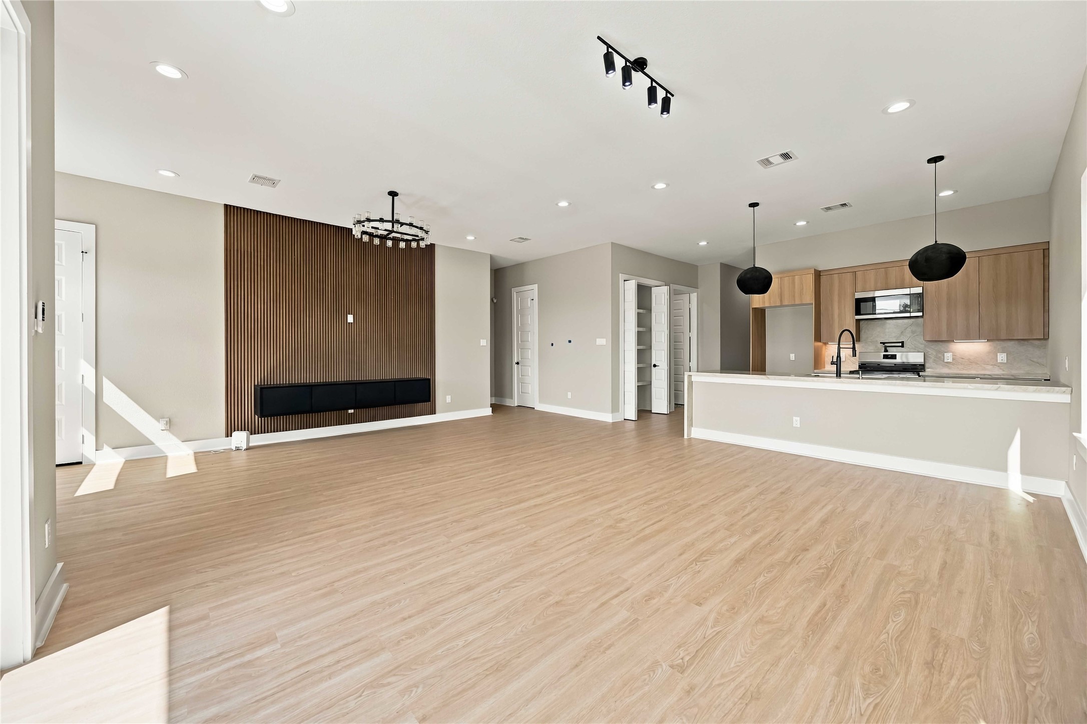 Open-concept living area featuring warm wood-look flooring, floating entertainment mantel, a striking vertical wood-slat accent wall, contemporary designer lighting, and a clean, modern aesthetic. The bright, airy layout flows seamlessly into the kitchen, creating an inviting space ideal for everyday living and entertaining.