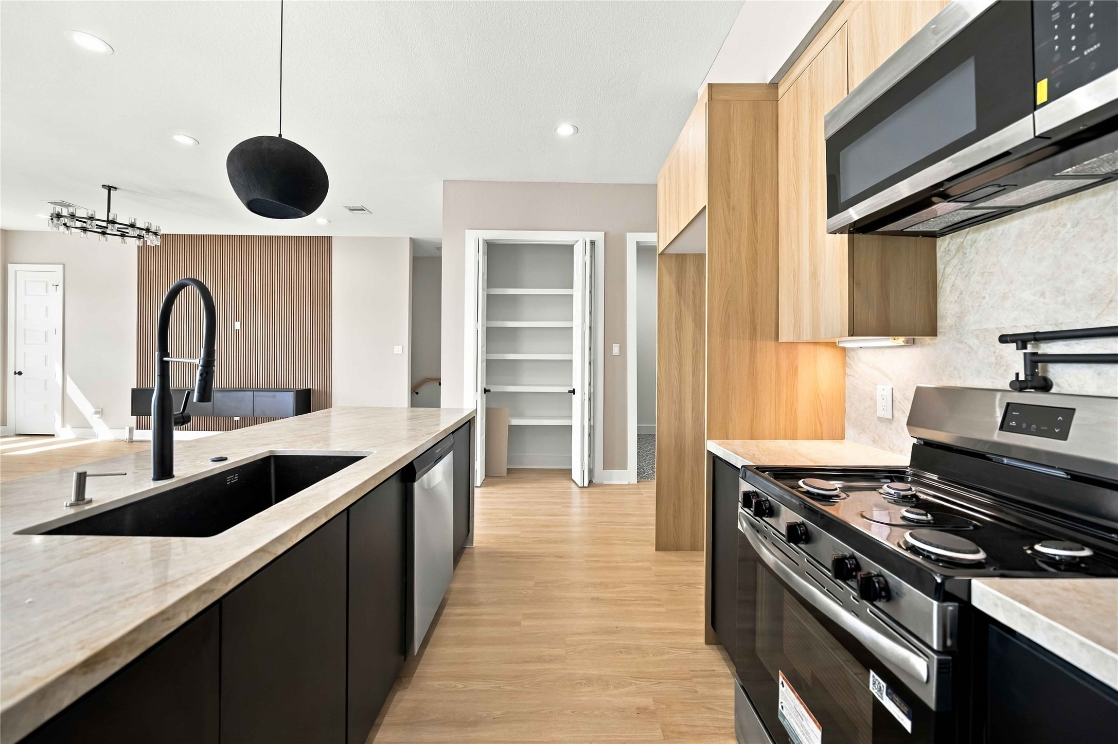 Sleek contemporary kitchen featuring warm natural wood cabinetry, contrasting black lower cabinets, elegant light stone countertops, a spacious island with a matte black gooseneck faucet, stainless steel appliances, and a modern backsplash. Recessed lighting and designer pendant fixtures complete this sophisticated, open-concept space.