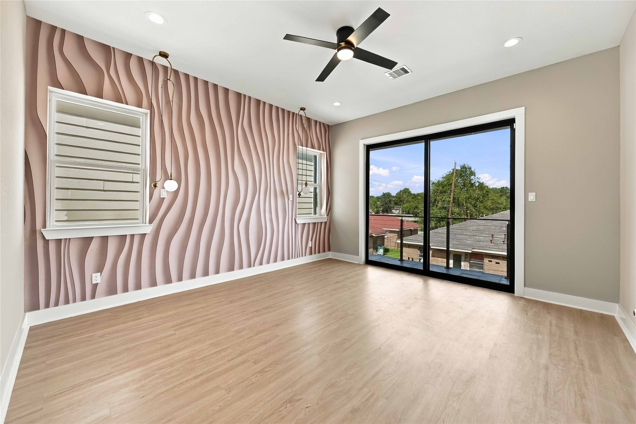 Contemporary primary bedroom highlighted by a distinctive sculptural blush-toned accent wall, warm wood-look flooring, modern pendant lighting, and a sleek ceiling fan. Expansive, black-framed sliding glass doors provide abundant natural light and access to a private balcony, creating a stylish and inviting retreat.
