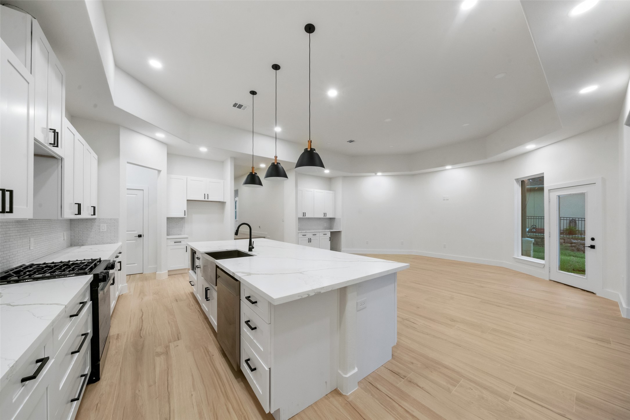 Beautifully designed gourmet kitchen featuring an oversized island with marble-look countertops, farmhouse-style sink, designer pendant lighting, and elegant white cabinetry with contrasting black hardware. The expansive open-concept layout flows effortlessly into the spacious living area, creating an exceptional setting for entertaining.