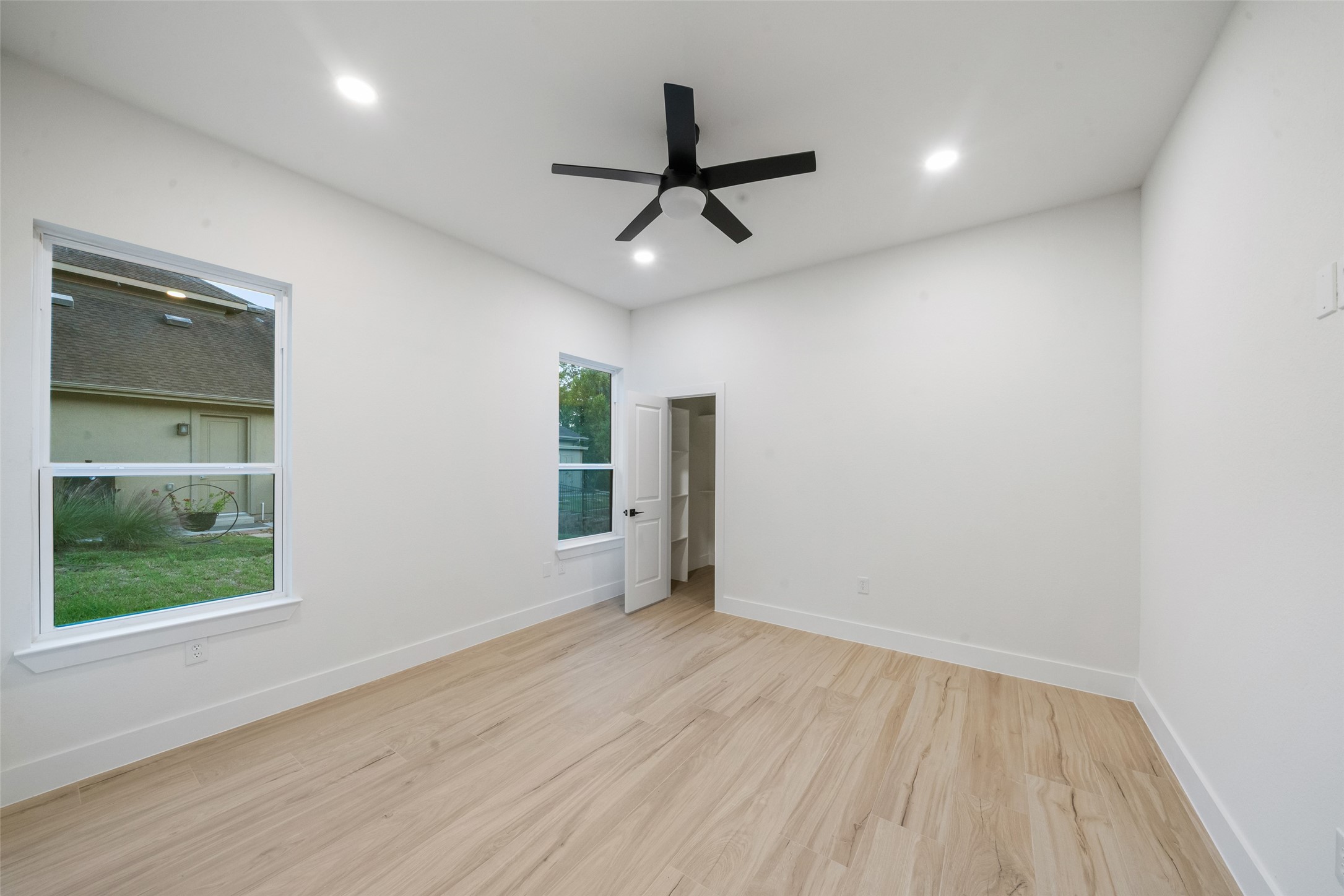 Spacious bedroom featuring beautiful wood-look flooring, a modern ceiling fan, recessed lighting, and large windows. Neutral finishes and an adjoining closet provide comfort and versatility.