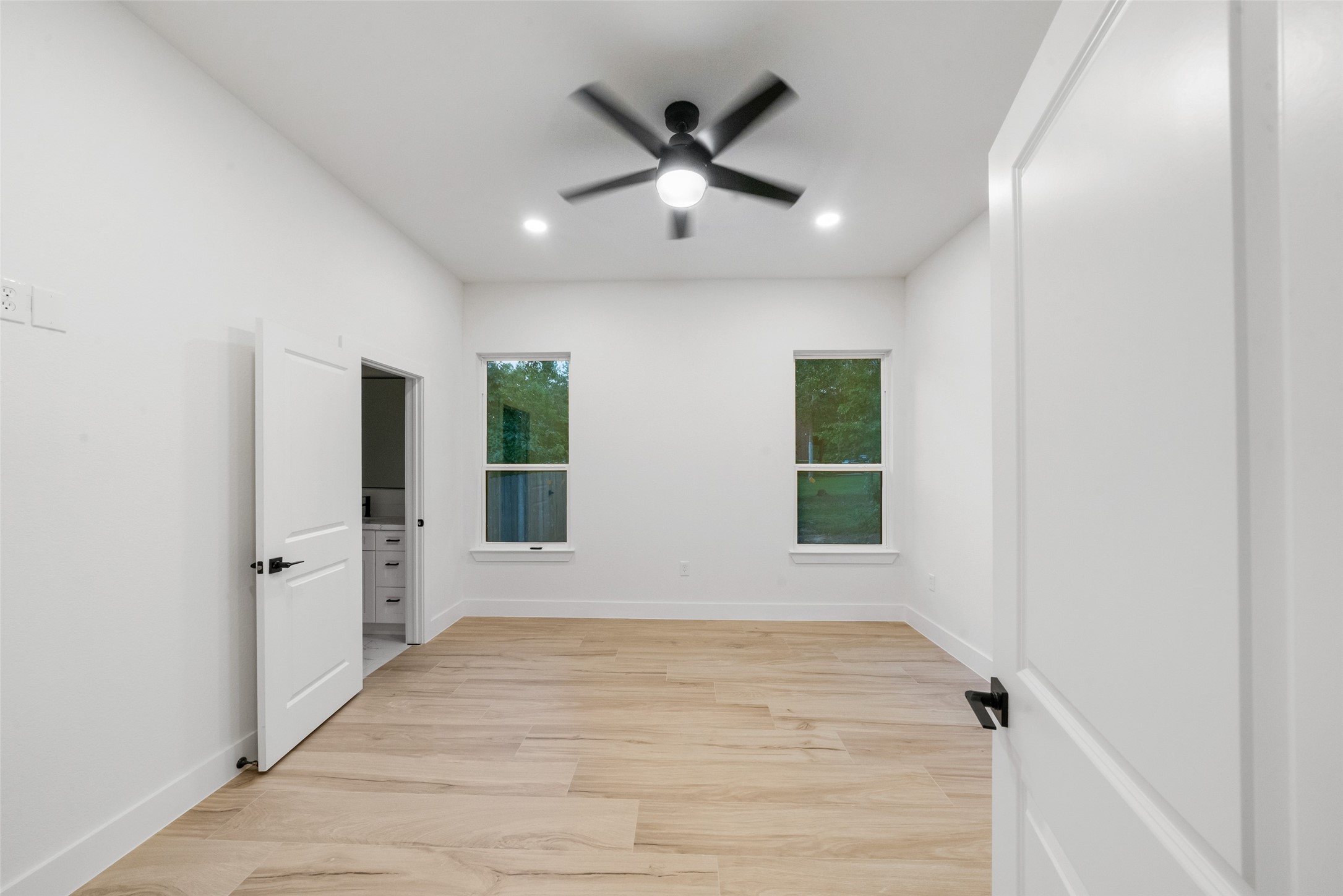 Bright and spacious bedroom featuring beautiful wood-look flooring, recessed lighting, a contemporary ceiling fan, and large windows overlooking the greenery. Neutral finishes and convenient access to an adjoining bathroom complete this inviting space.
