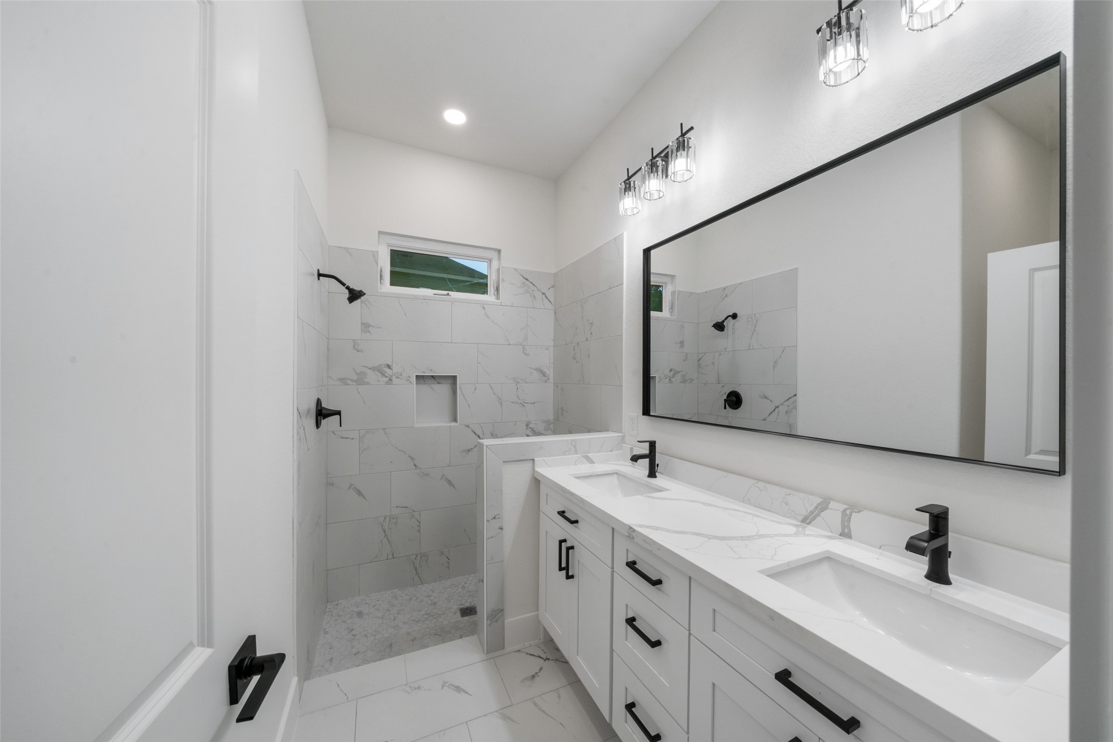 Beautifully appointed bathroom featuring a spacious dual-sink vanity, marble-look countertops, sleek black fixtures, and a tiled walk-in shower with built-in niche. Elegant finishes create a sophisticated, spa-inspired atmosphere.
