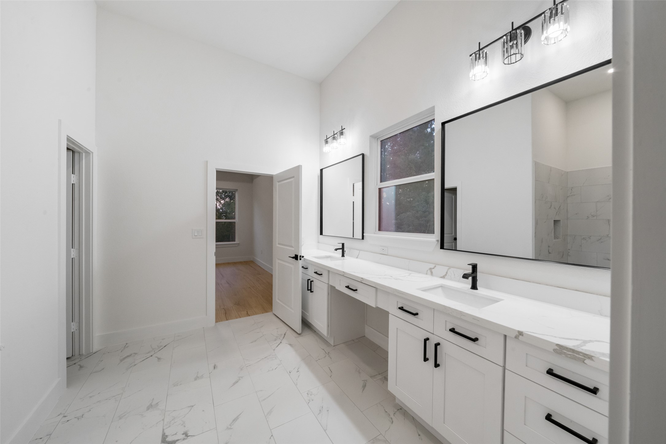 Luxurious primary bathroom showcasing an extended dual-sink vanity with elegant countertops, individual framed mirrors, and sophisticated black fixtures. A large window welcomes natural light, beautifully complementing the room's bright, timeless finishes.