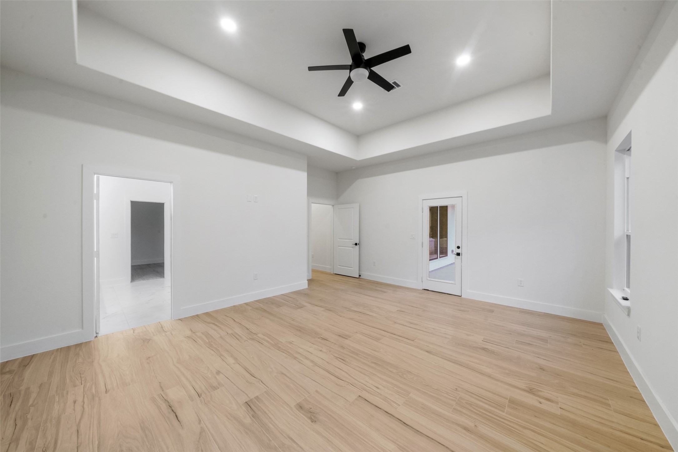 Impressive primary suite featuring soaring tray ceilings, recessed lighting, modern ceiling fan, and beautiful wood-look flooring. An expansive layout provides flexibility for furnishings, with convenient access to the primary bath and covered outdoor living area.
