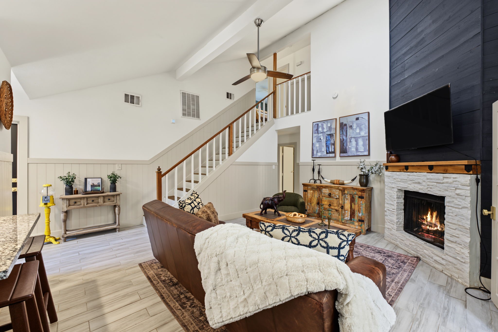 Soaring vaulted living room boasts a wood beam, floor-to-ceiling black shiplap and a white stacked stone fireplace with rustic wood mantel!