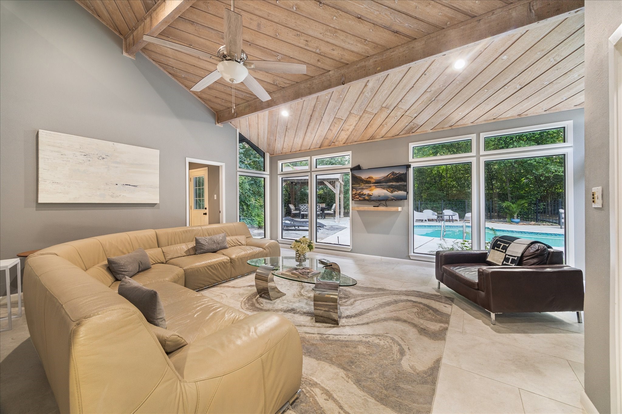 Another spacious living room features a vaulted wooden ceiling and large windows offering a view of the pool and lush greenery outside.