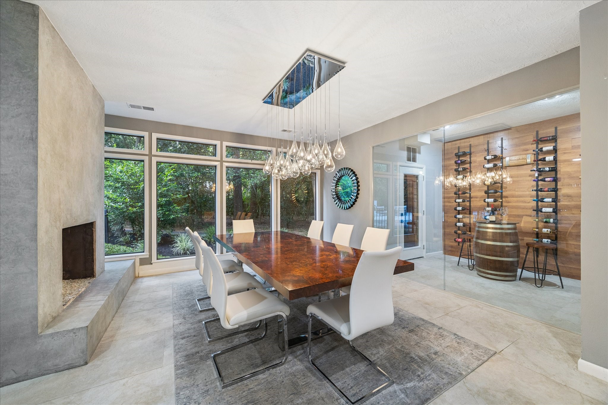 The dining room features a large wooden table with sleek white chairs, a stylish light fixture, and expansive windows offering natural views. The room includes a contemporary fireplace and a glass-enclosed wine display area, creating a luxurious and inviting atmosphere.