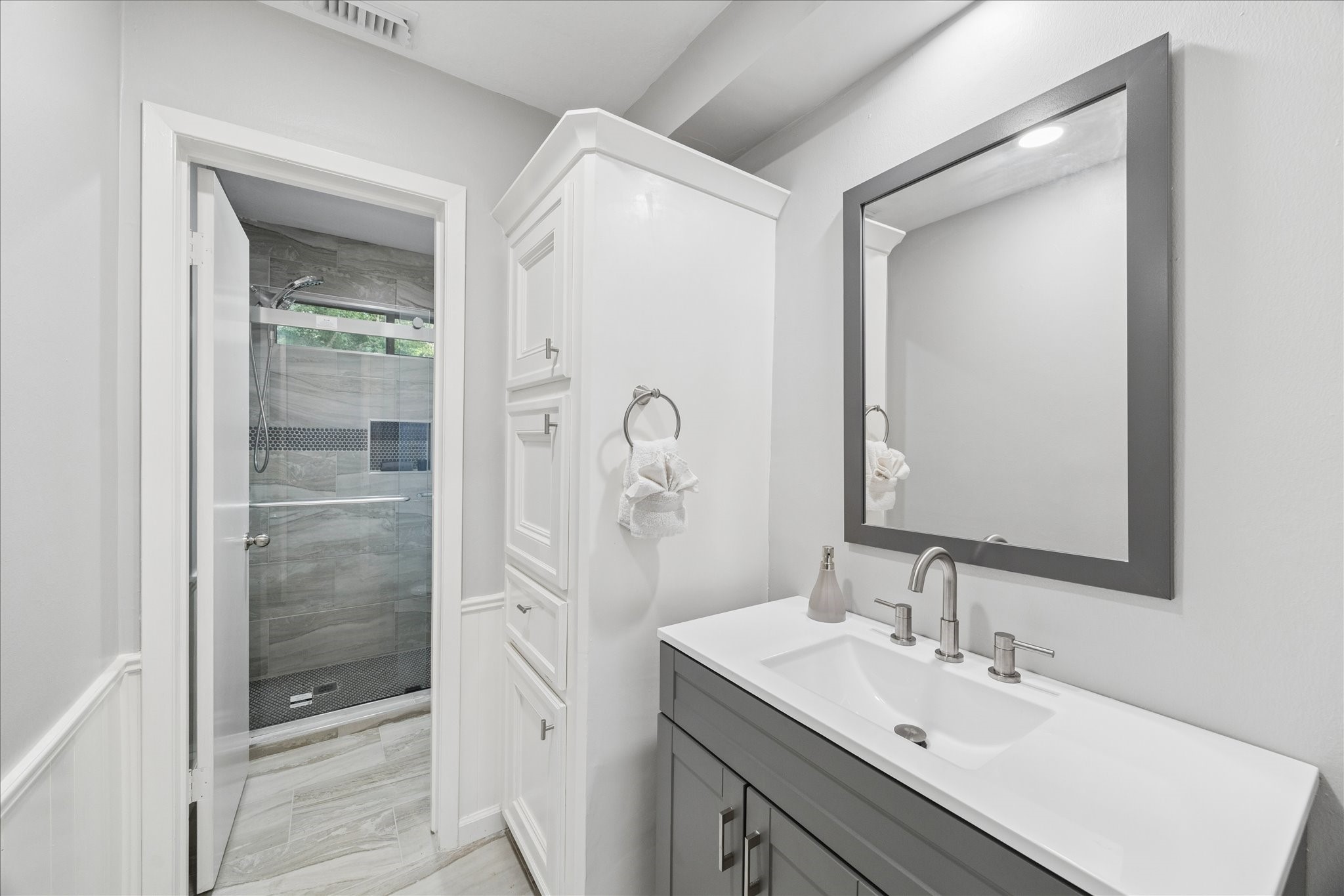 This bathroom features a modern design with a sleek vanity and large mirror. It includes a spacious glass-enclosed shower with stylish tile work. The room offers ample storage and a clean, bright aesthetic.