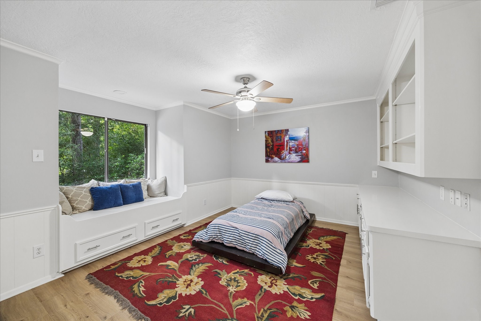 The 2nd-floor bedroom features a built-in window seat with storage, a ceiling fan, and a comfortable bed. The neutral walls and wood flooring create a warm atmosphere, complemented by a vibrant floral rug. Built-in shelves and a desk area offer ample storage and workspace.