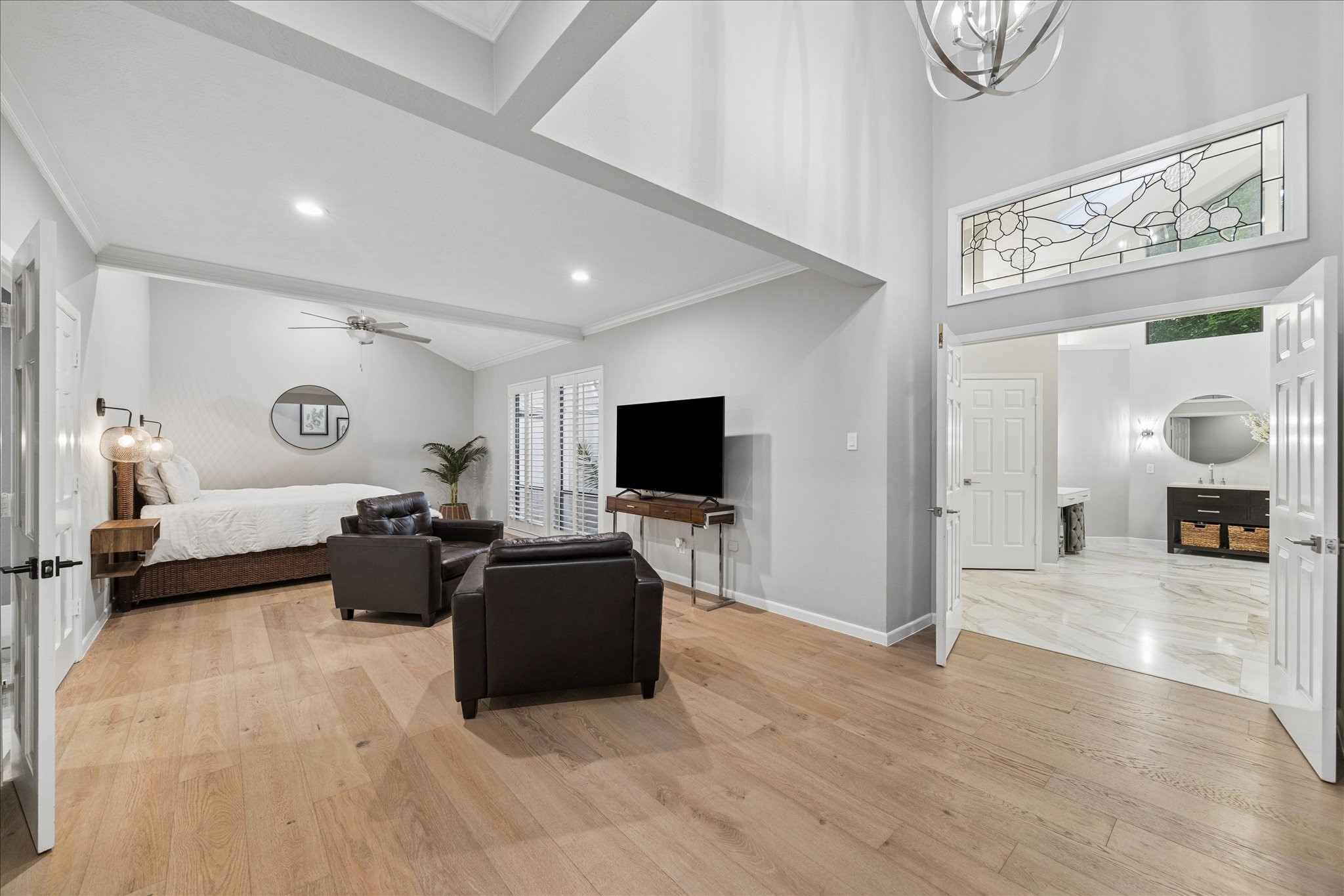 The primary suite features vaulted ceilings and wood flooring. It features a cozy seating area, large windows with plantation shutters, and an adjoining luxurious bathroom with marble flooring. The room is elegantly lit, enhancing its airy and inviting atmosphere.