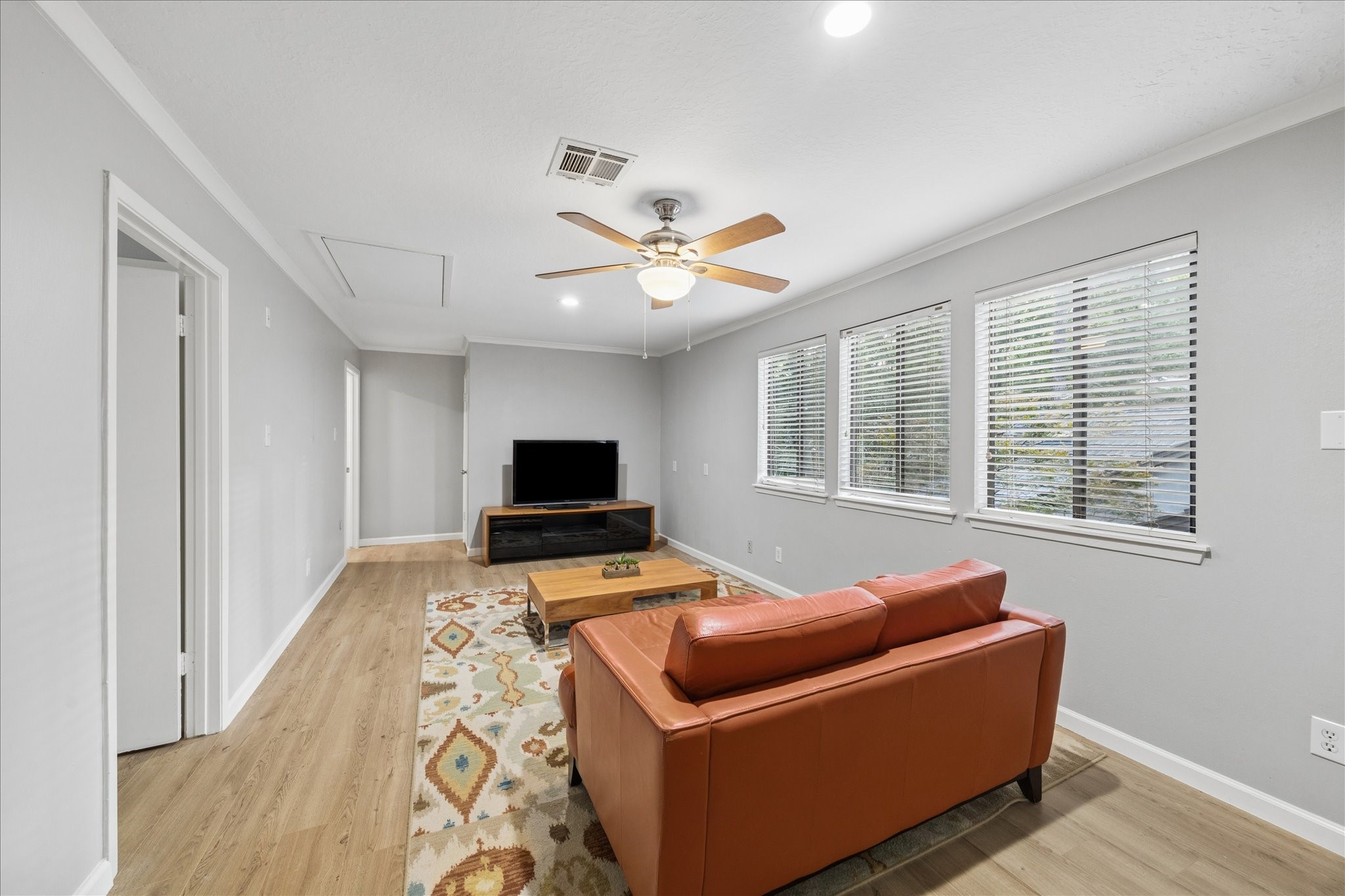 This cozy 2nd-floor living room features a modern ceiling fan, light gray walls, and wood flooring. It includes a comfortable sofa, a stylish area rug, and a TV setup. Large windows provide ample natural light, creating a bright and inviting atmosphere.