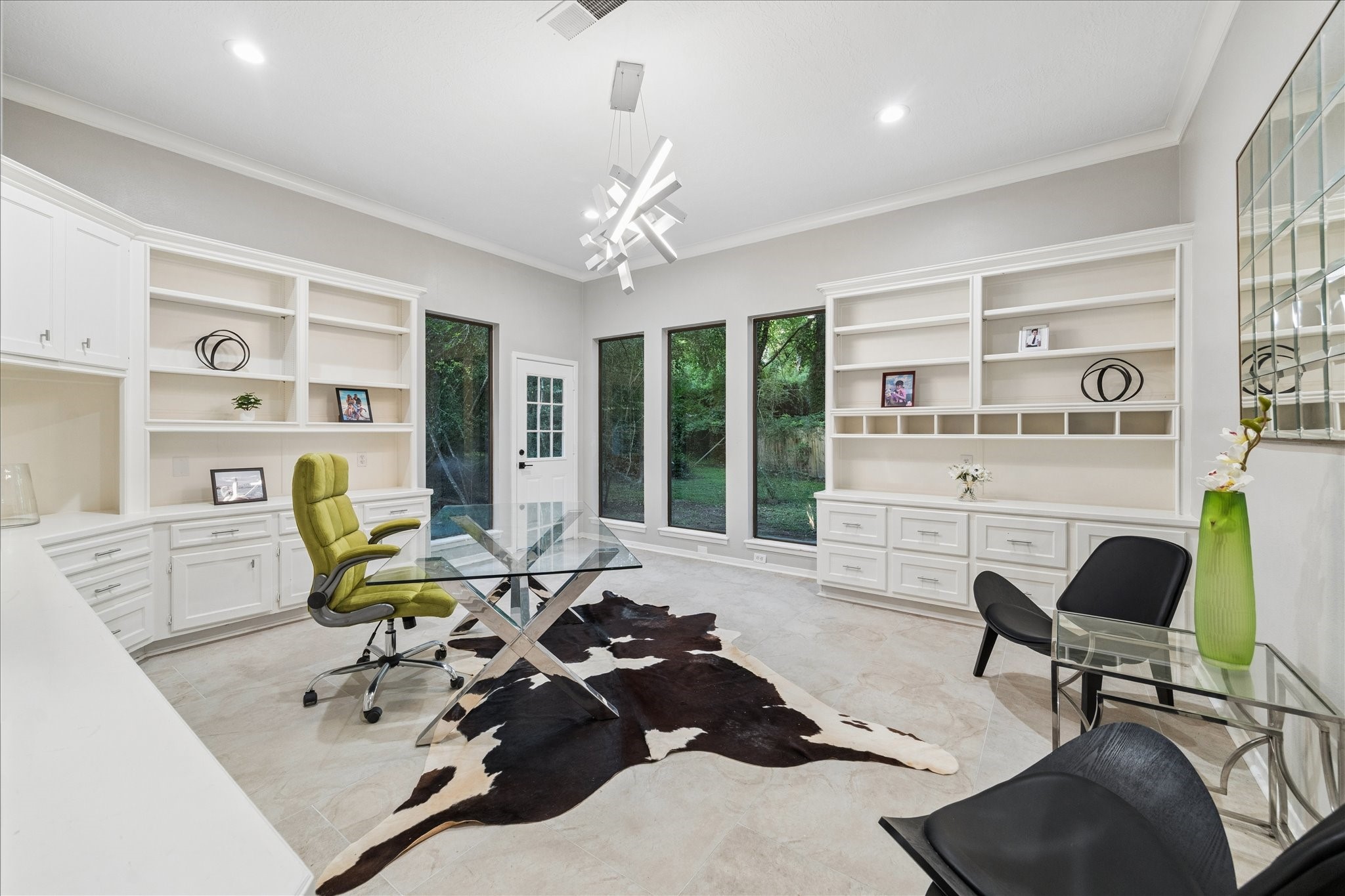 This home office features sleek modern design with ample natural light from large windows. It includes built-in shelving, a stylish glass desk, and contemporary lighting, creating a functional and elegant workspace.